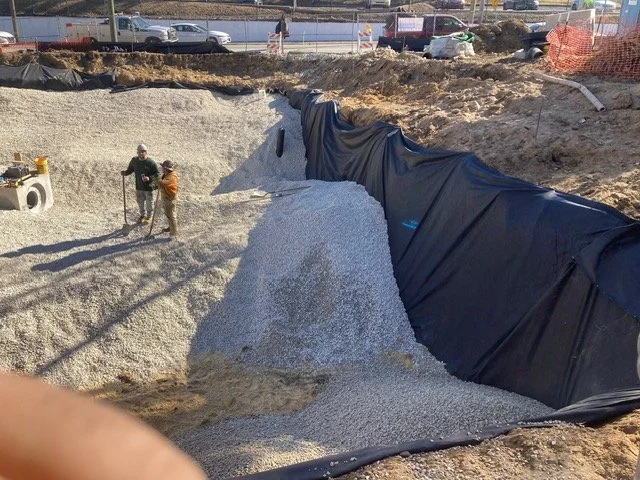 Construction workers working in a large excavation site with a pile of gravel and black plastic lining the sides.