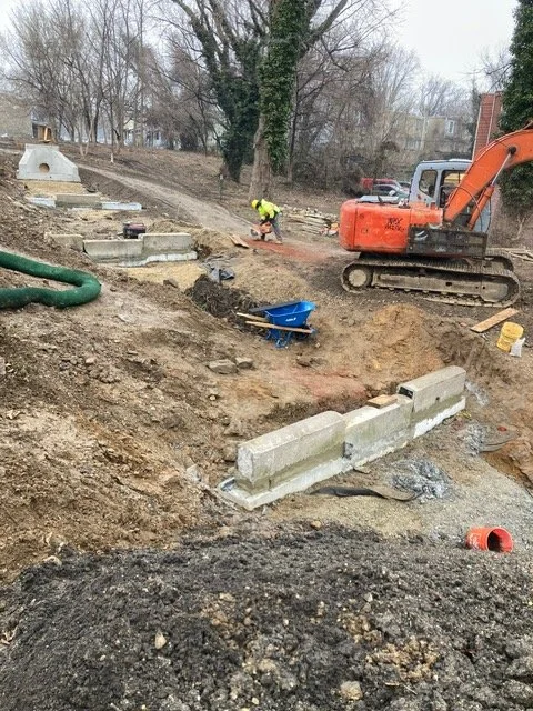 Construction site with an orange excavator and a worker in a yellow safety vest installing concrete blocks for a wall.
