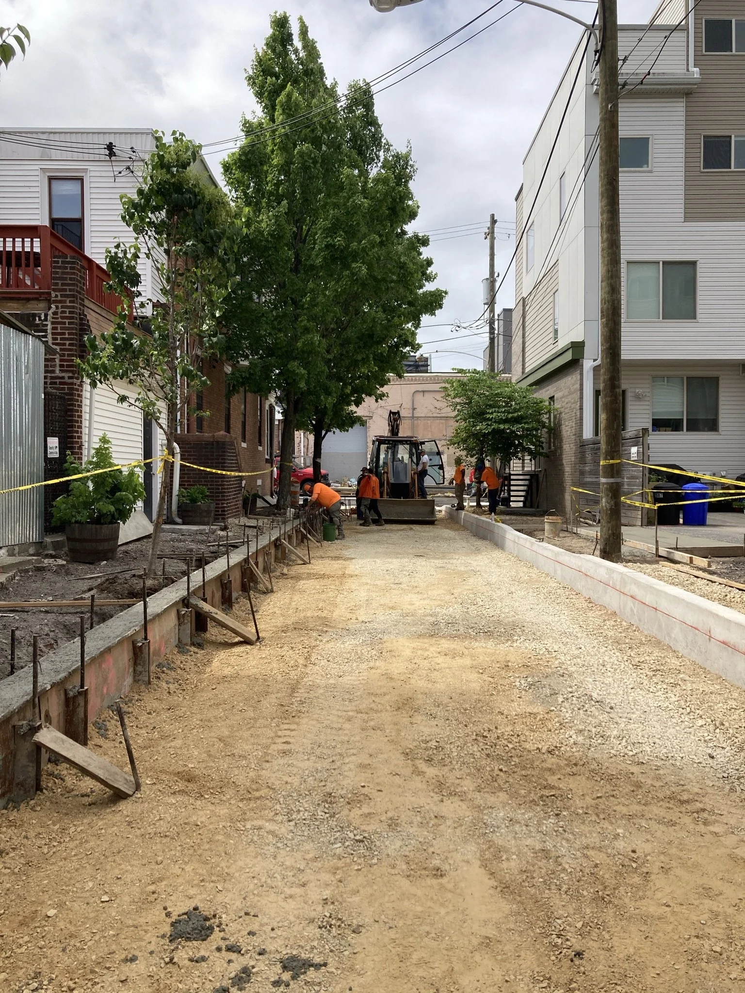 Construction workers working on a sidewalk renovation in an urban neighborhood with multi-story residential buildings, trees, and utility poles.