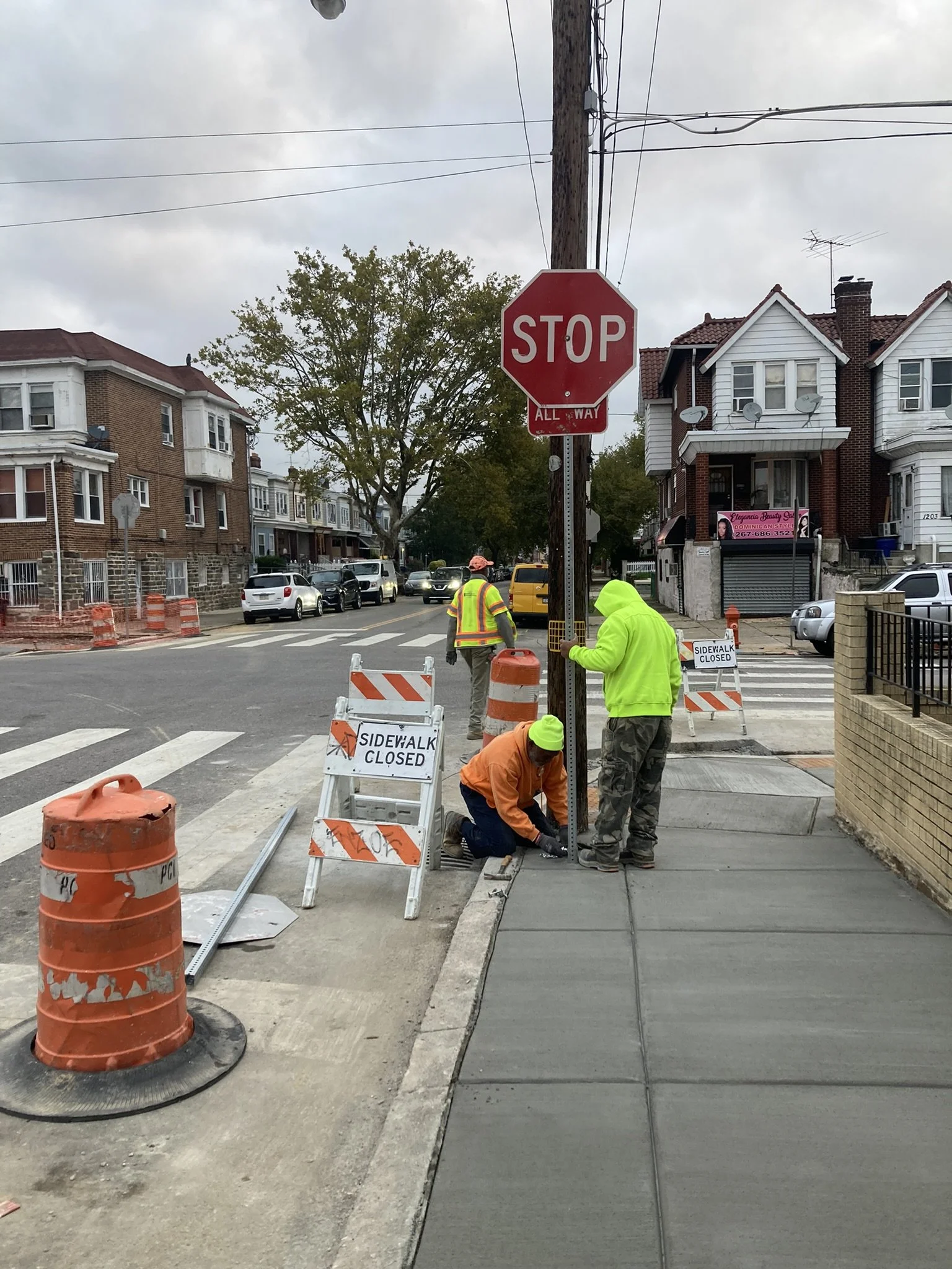 Construction workers are installing a stop sign on a city street, with street and sidewalk work in progress and traffic cones and signs indicating sidewalk is closed.