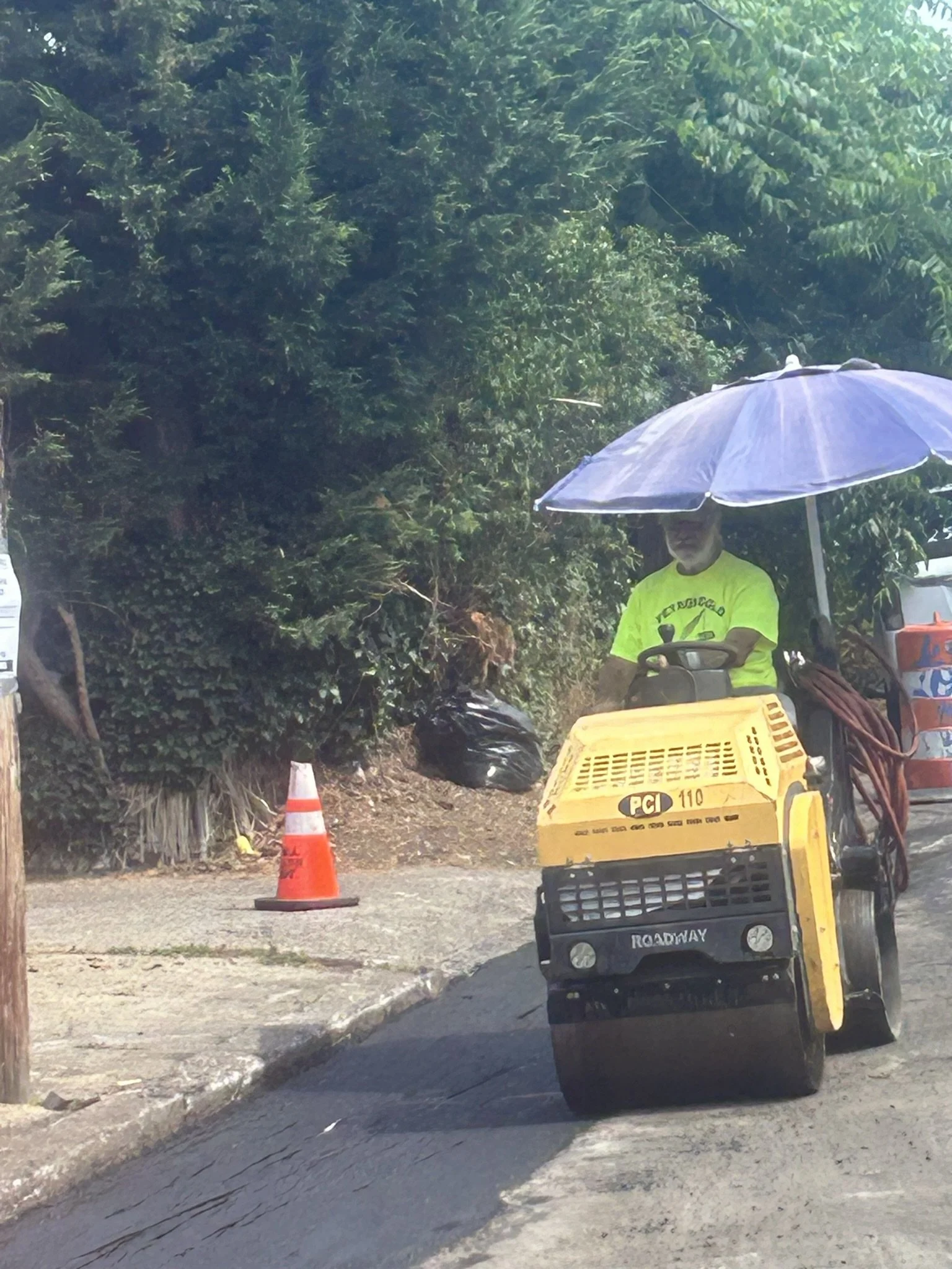 A person operating road construction equipment on a street, holding an umbrella for shade, with trees on the side and traffic cones nearby.