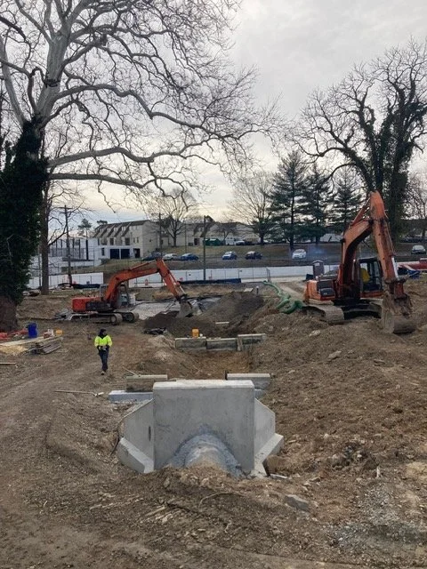 Construction site with two excavators digging into the ground, a worker in a yellow vest, concrete pipe, and construction materials, with leafless trees and a road in the background.