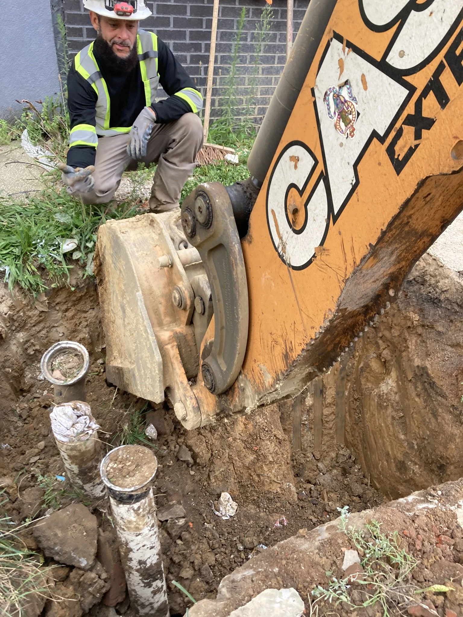 A construction worker crouching near an excavator bucket that is digging into the ground, with pipes and soil visible in the hole.