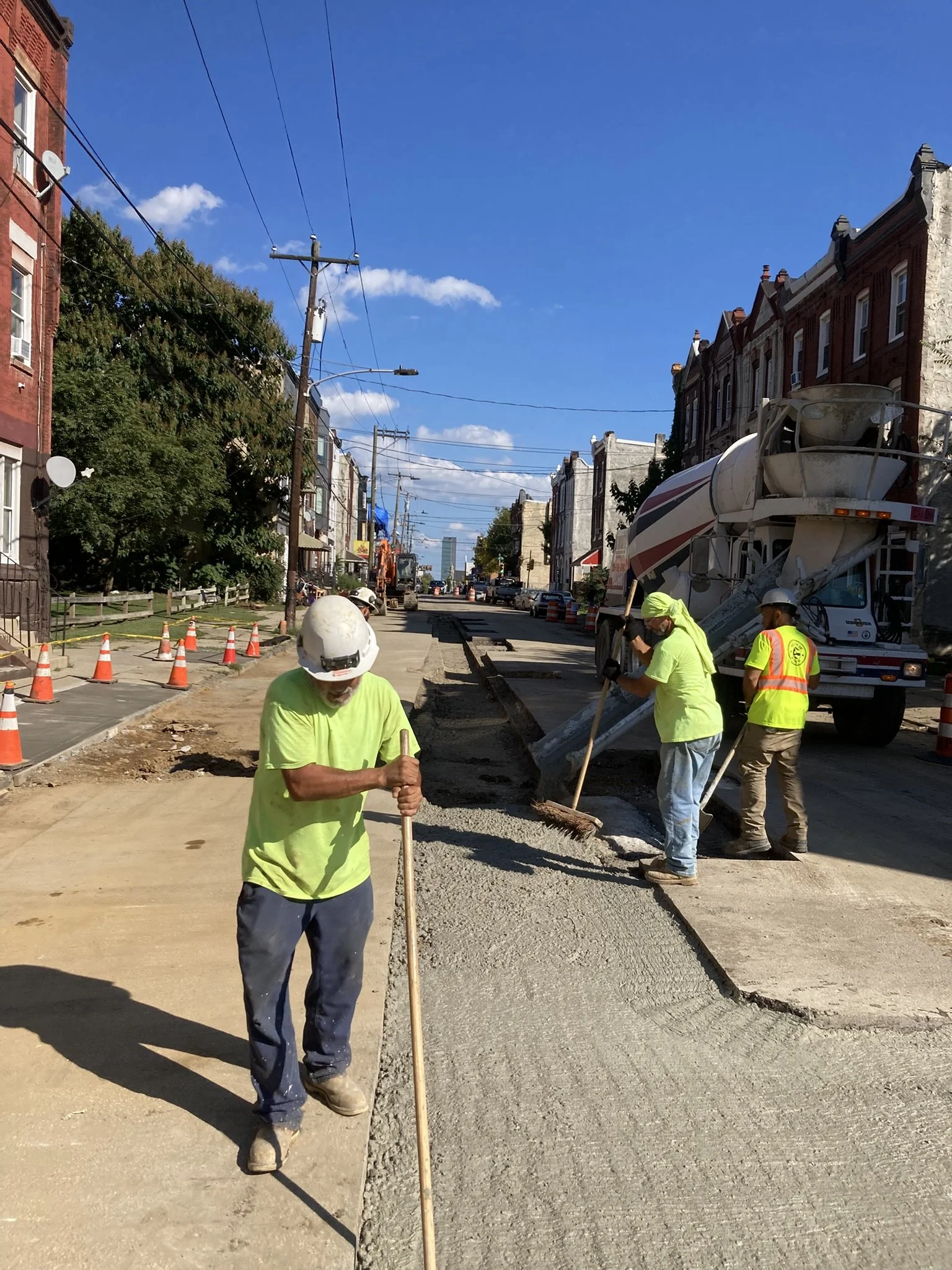 Construction workers in yellow safety vests and helmets working on a street with asphalt and concrete, with a cement mixer truck in the background and orange traffic cones along the sidewalk, under a clear blue sky.