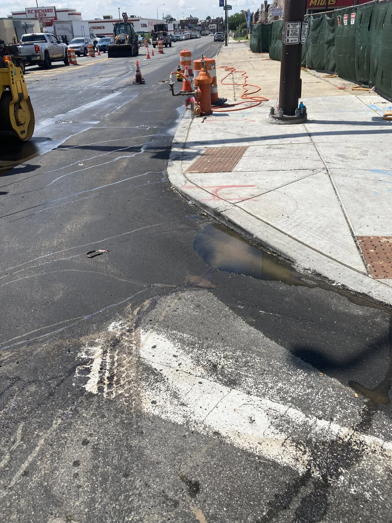 Street with ongoing road work, construction cones, and machinery; wet asphalt and water puddles, sidewalk with construction barriers, utility pole, and traffic in the background.