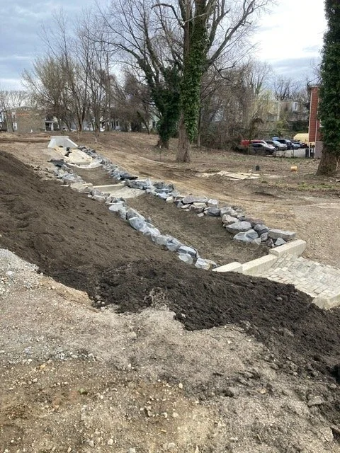 Landscape with a newly constructed stone-lined pathway on an outdoor area, with trees, parked cars, and houses in the background.