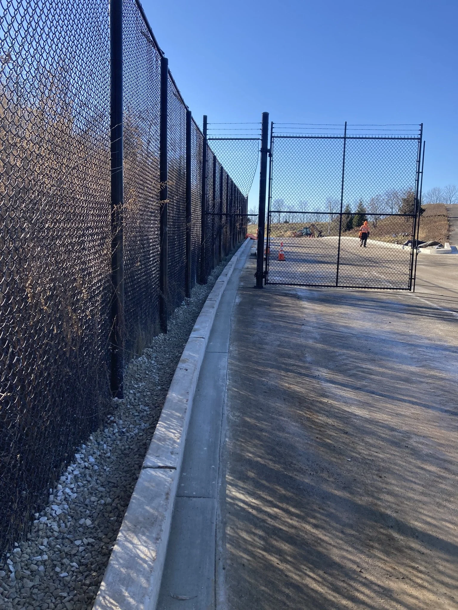 View of a new chain-link fence and gate at a construction site with a worker in the background under a clear blue sky.