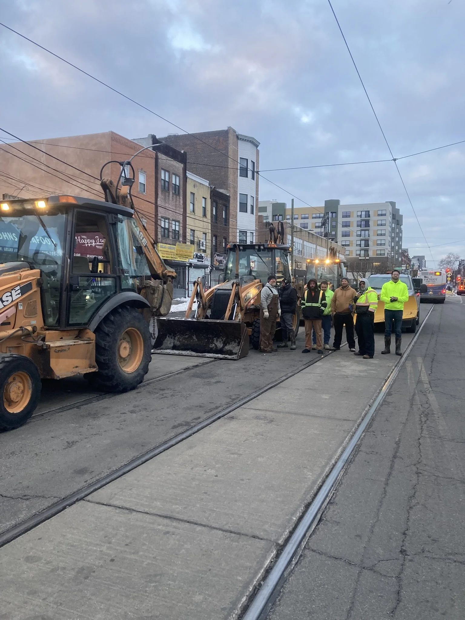 City street scene with construction equipment and workers gathered around a snow plow on tram tracks, with residential buildings and overcast sky in the background.