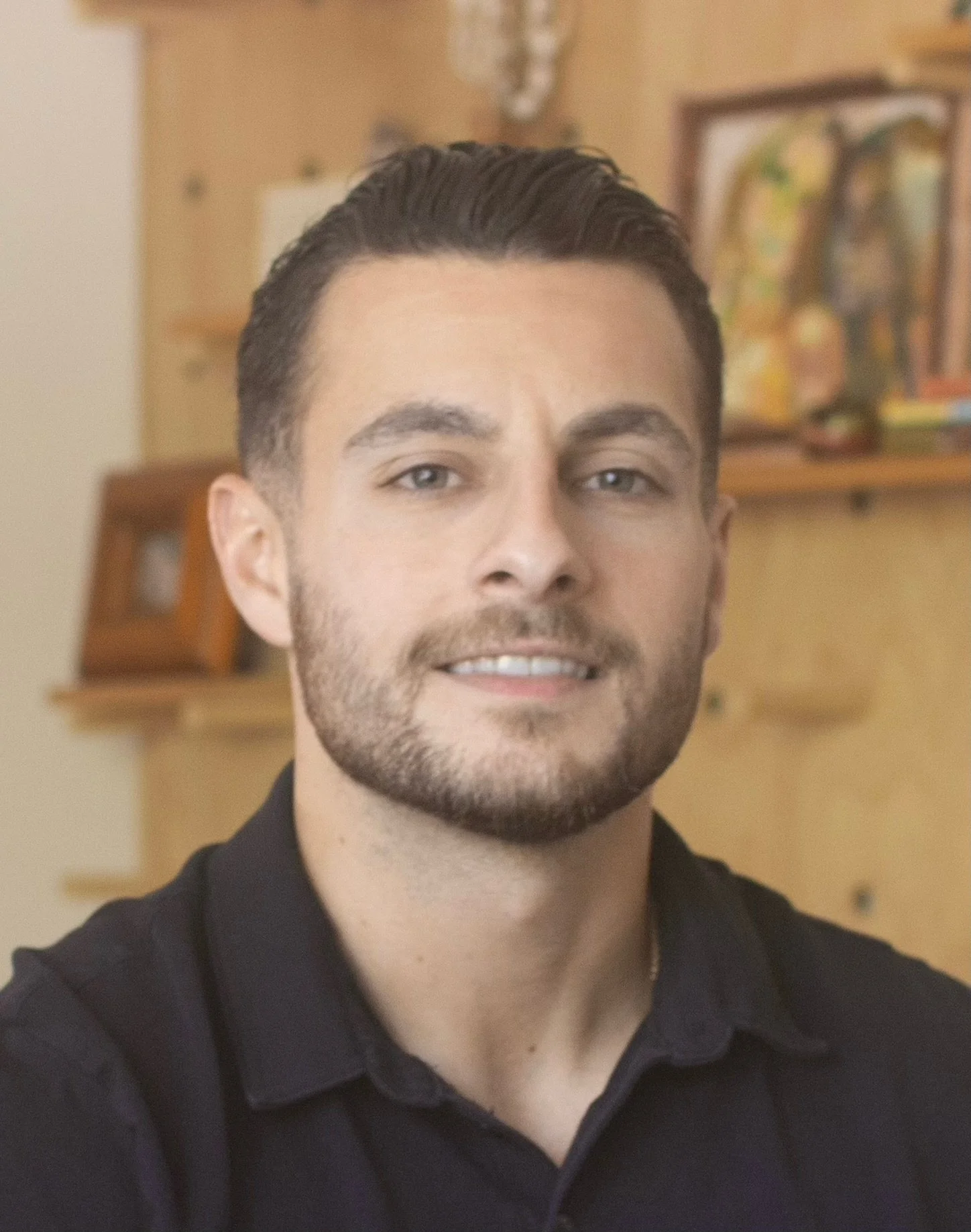 A young man with dark hair and a beard wearing a black collared shirt, smiling slightly, standing in an indoor setting with blurred wall decorations and furniture in the background.