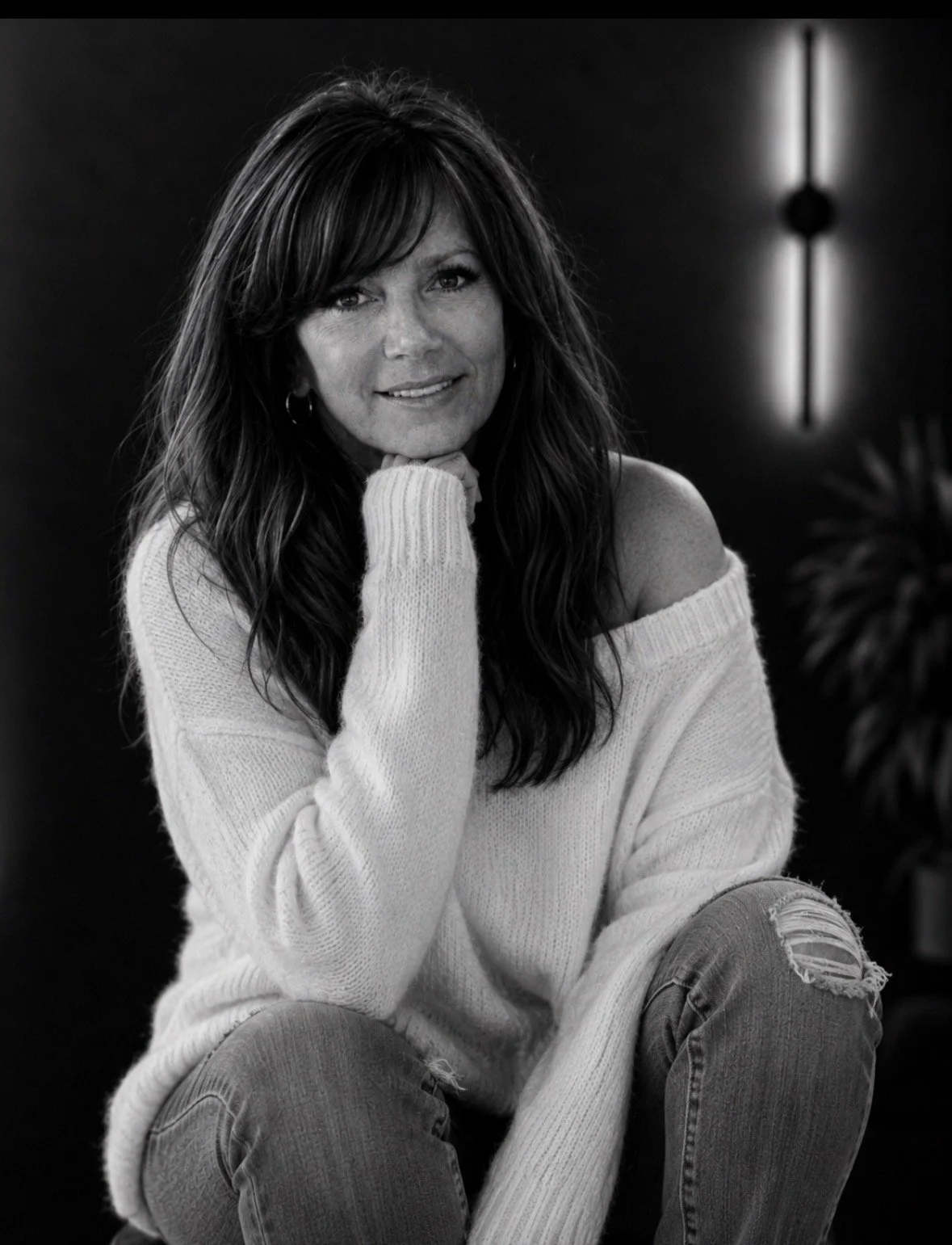 A woman with long dark hair and wearing a loose off-shoulder white sweater, sitting down with her chin resting on her hand, in a dimly lit room.