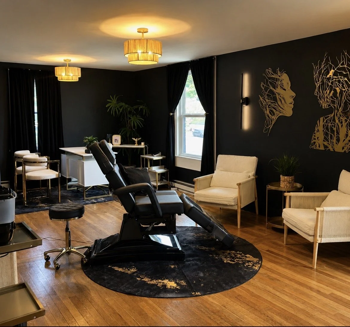 A modern, stylish medical or beauty treatment room featuring a black reclining chair, plush seating, plants, wall art, and warm lighting.