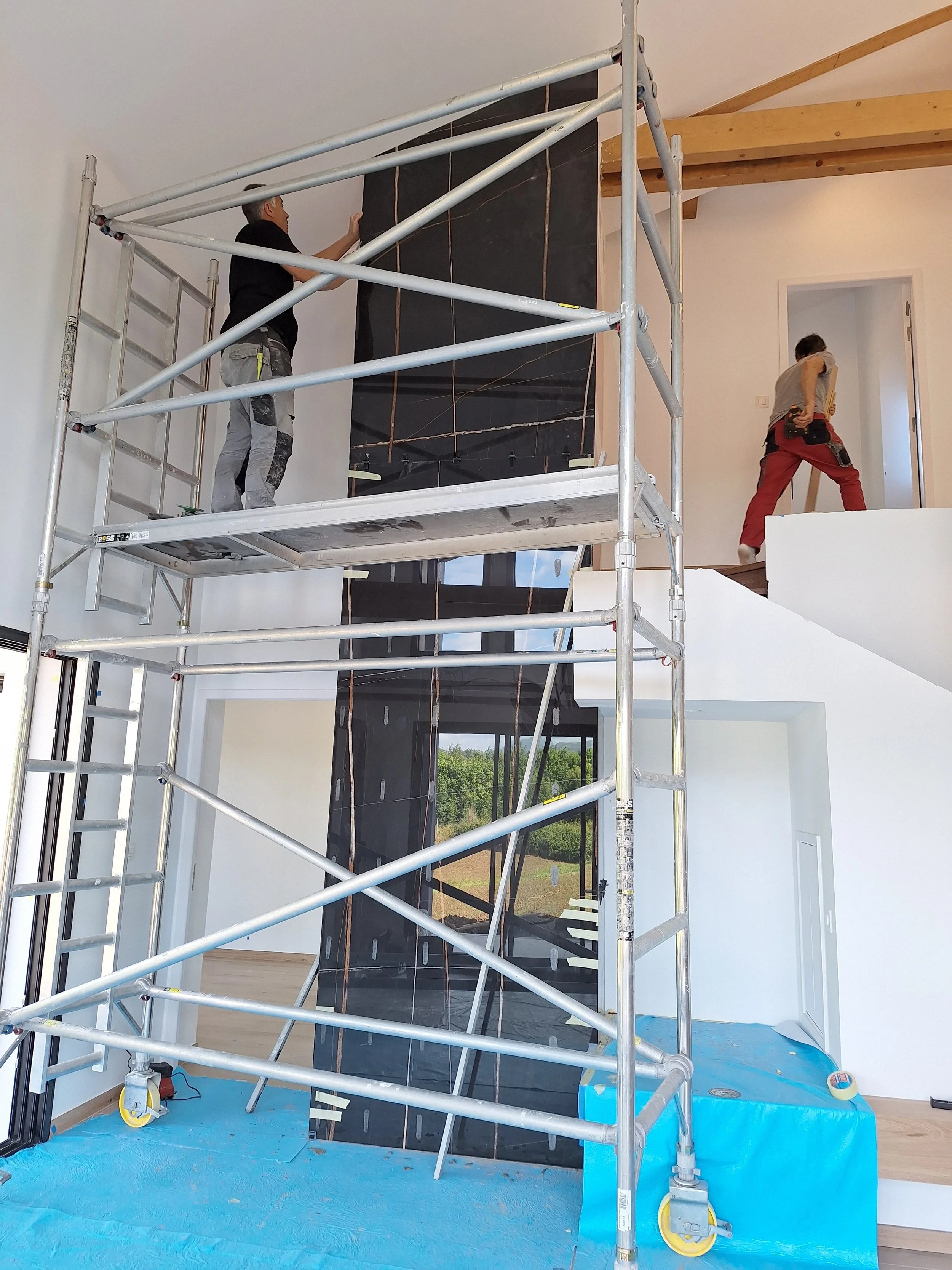 Deux ouvriers effectuent des travaux d'aménagement intérieur dans une maison en utilisant une échafaude en métal, près d'une grande cheminée noire, dans une pièce spacieuse avec murs blancs et plafond en bois.