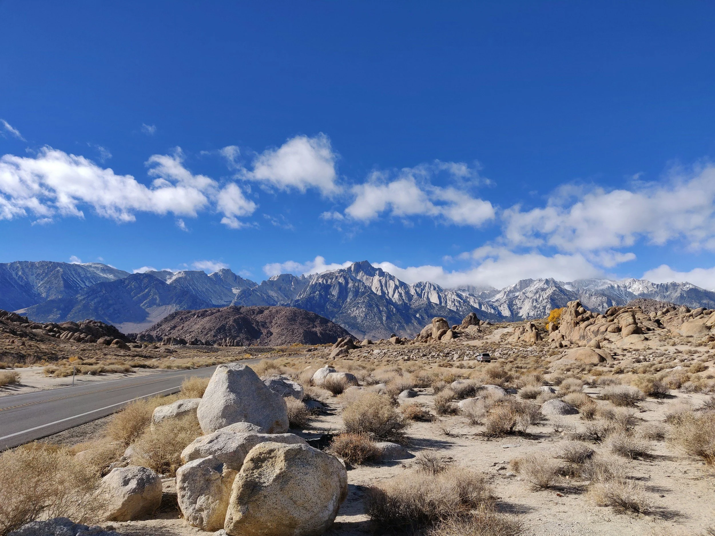 Scenic view of a desert landscape with large rocks, dry shrubs, a paved road, and snow-capped mountains under a blue sky with scattered clouds.