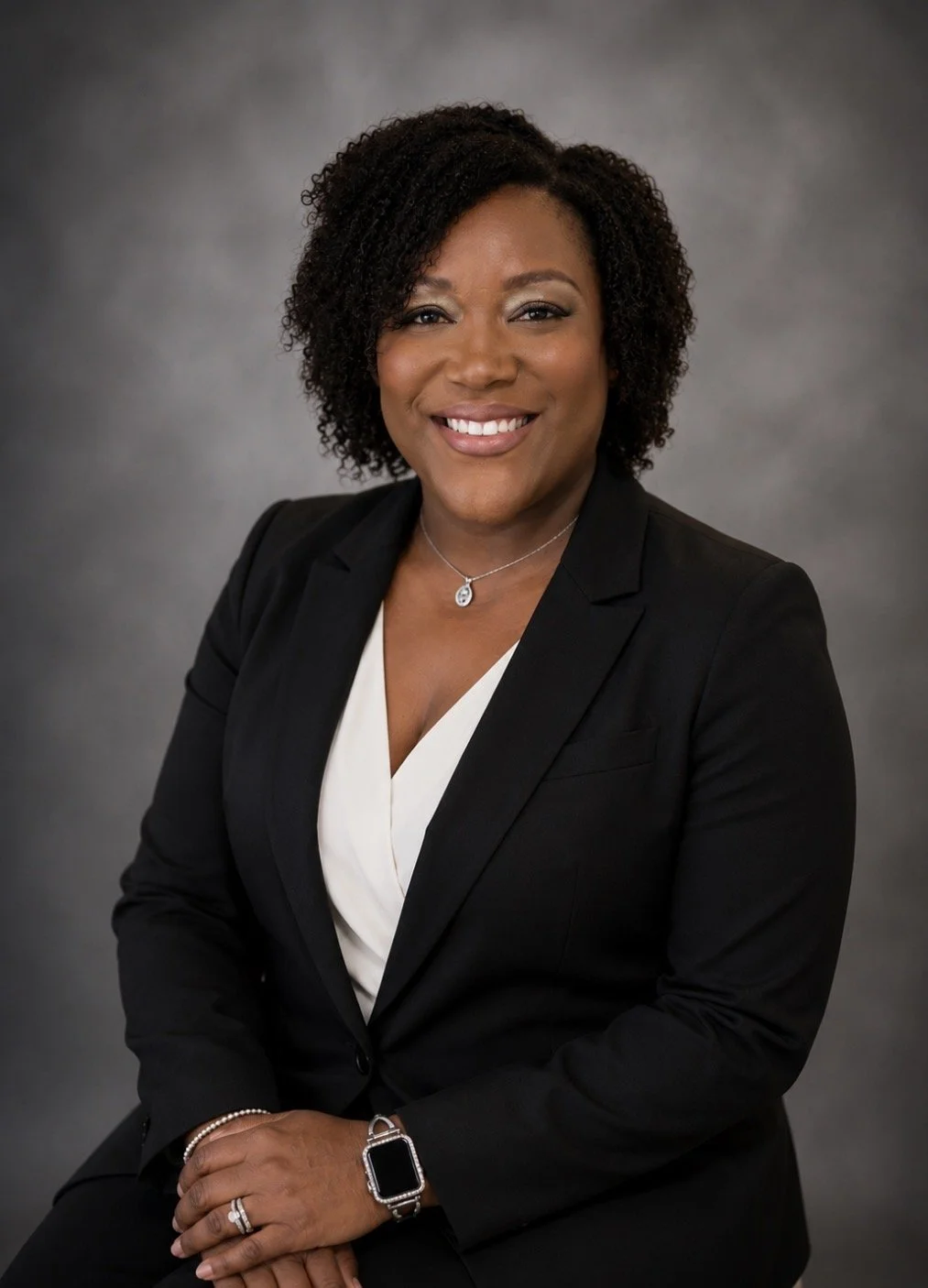 Professional woman with curly hair, wearing a black blazer and white blouse, smiling, sitting with her hands clasped.