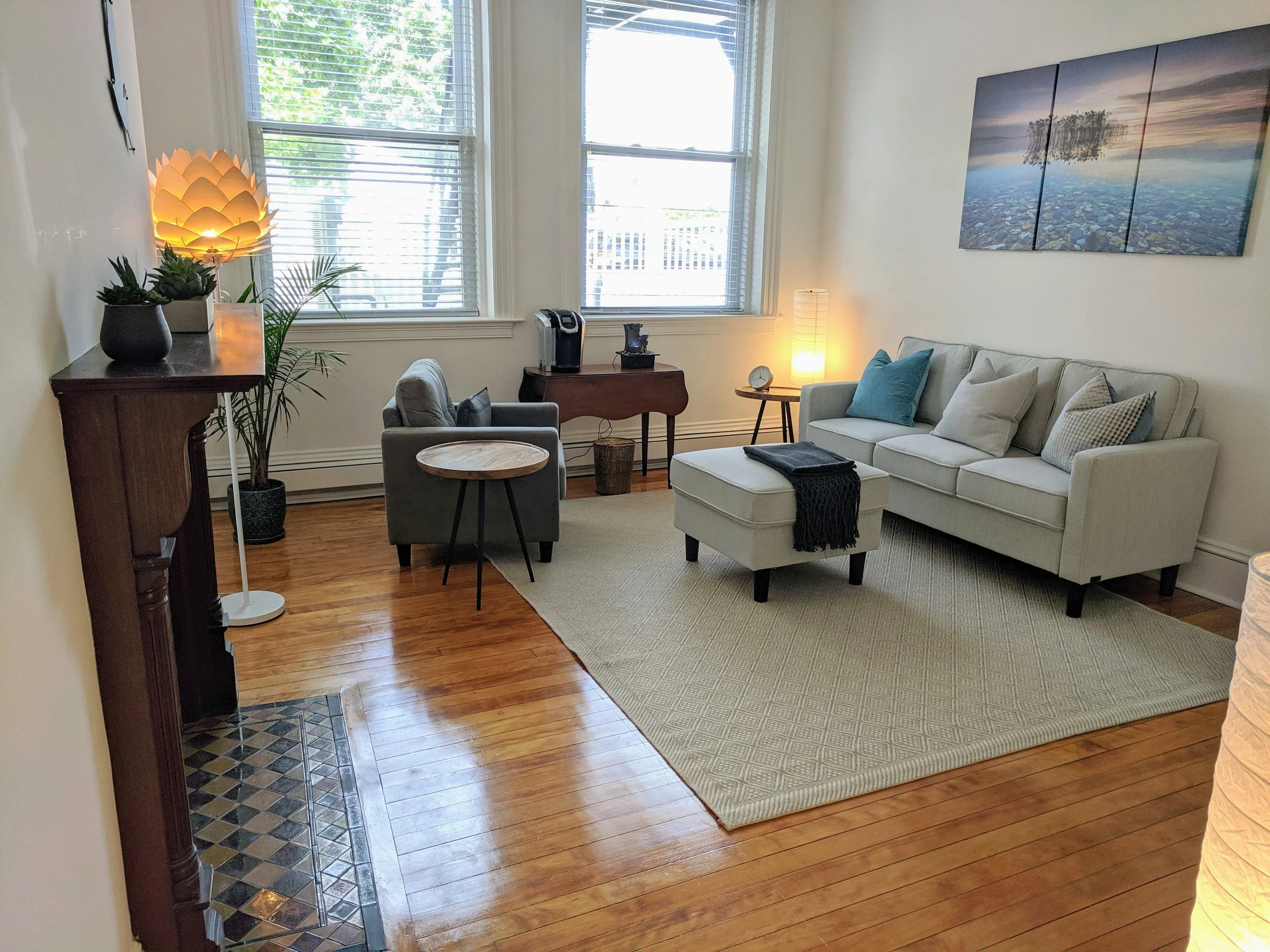 Living room with hardwood floors, beige sofa with four pillows, a small ottoman, a grey armchair, a round side table, a wooden sideboard, a tall lamp, plants, and a large landscape painting on the wall.