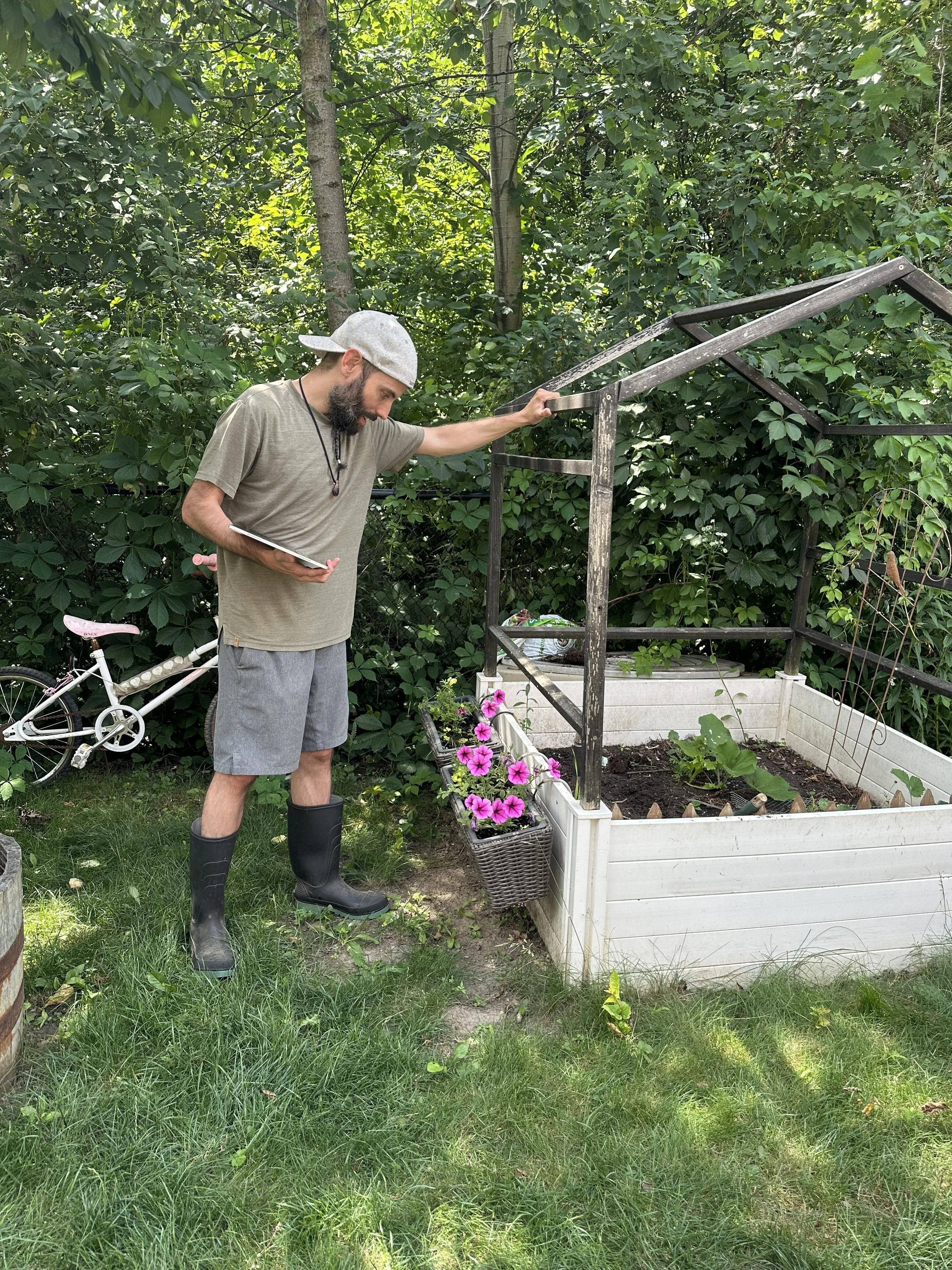 A man wearing a gray cap, t-shirt, shorts, and rubber boots is looking at a tablet while standing next to a raised garden bed with some plants. There is a basket of pink flowers hanging on the side of the garden bed, and a bicycle leaning against the