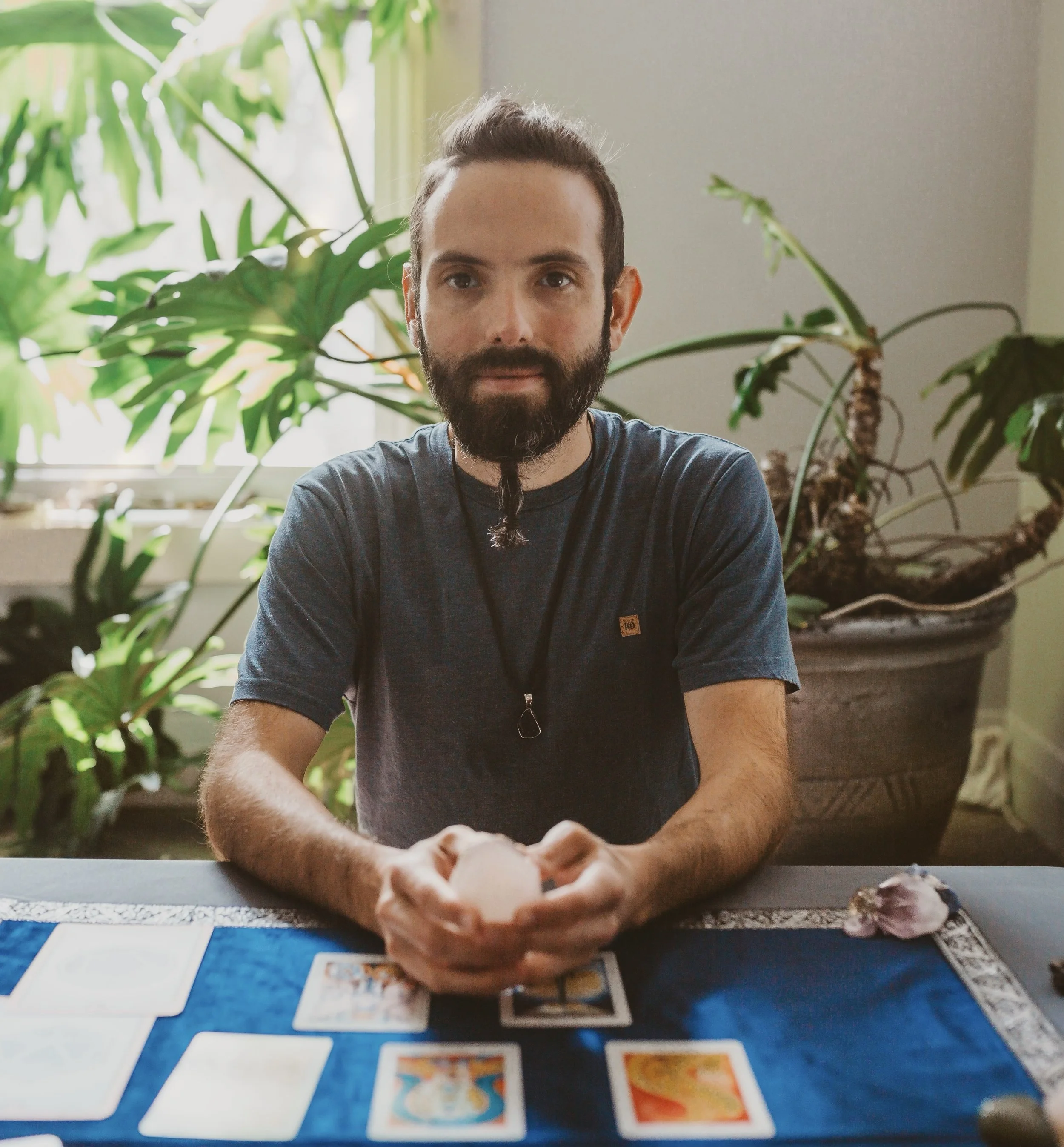 A man with a beard and long hair styled in a bun, sitting at a table with tarot cards, holding a pink crystal, with large green houseplants in the background.