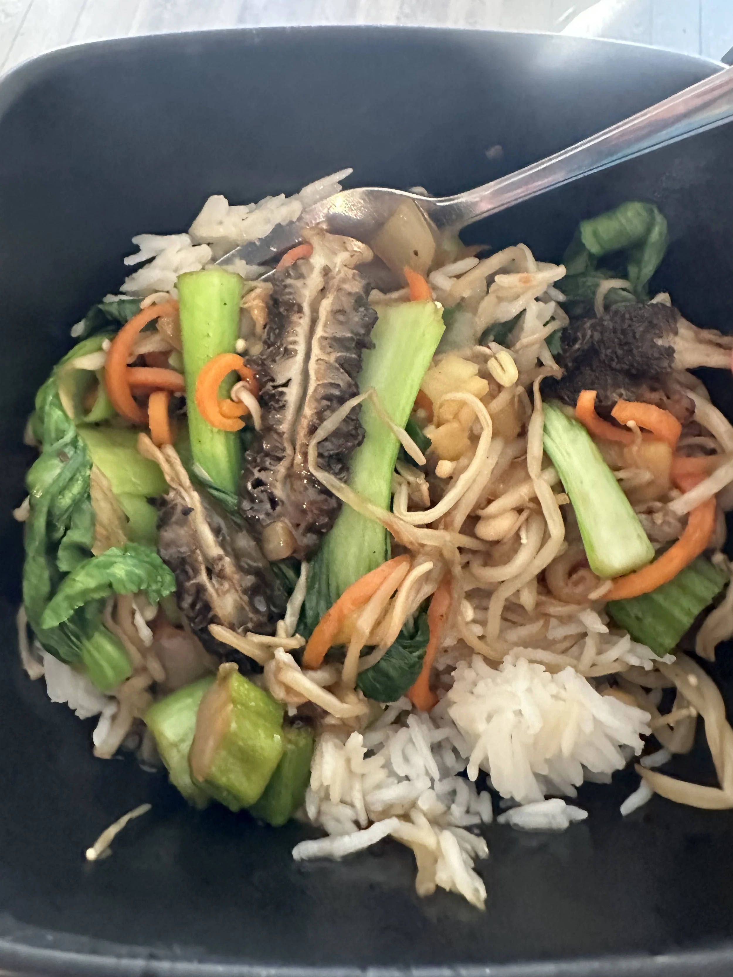 Stir-fried vegetables with rice in a black bowl, including okra, bean sprouts, carrots, and leafy greens.