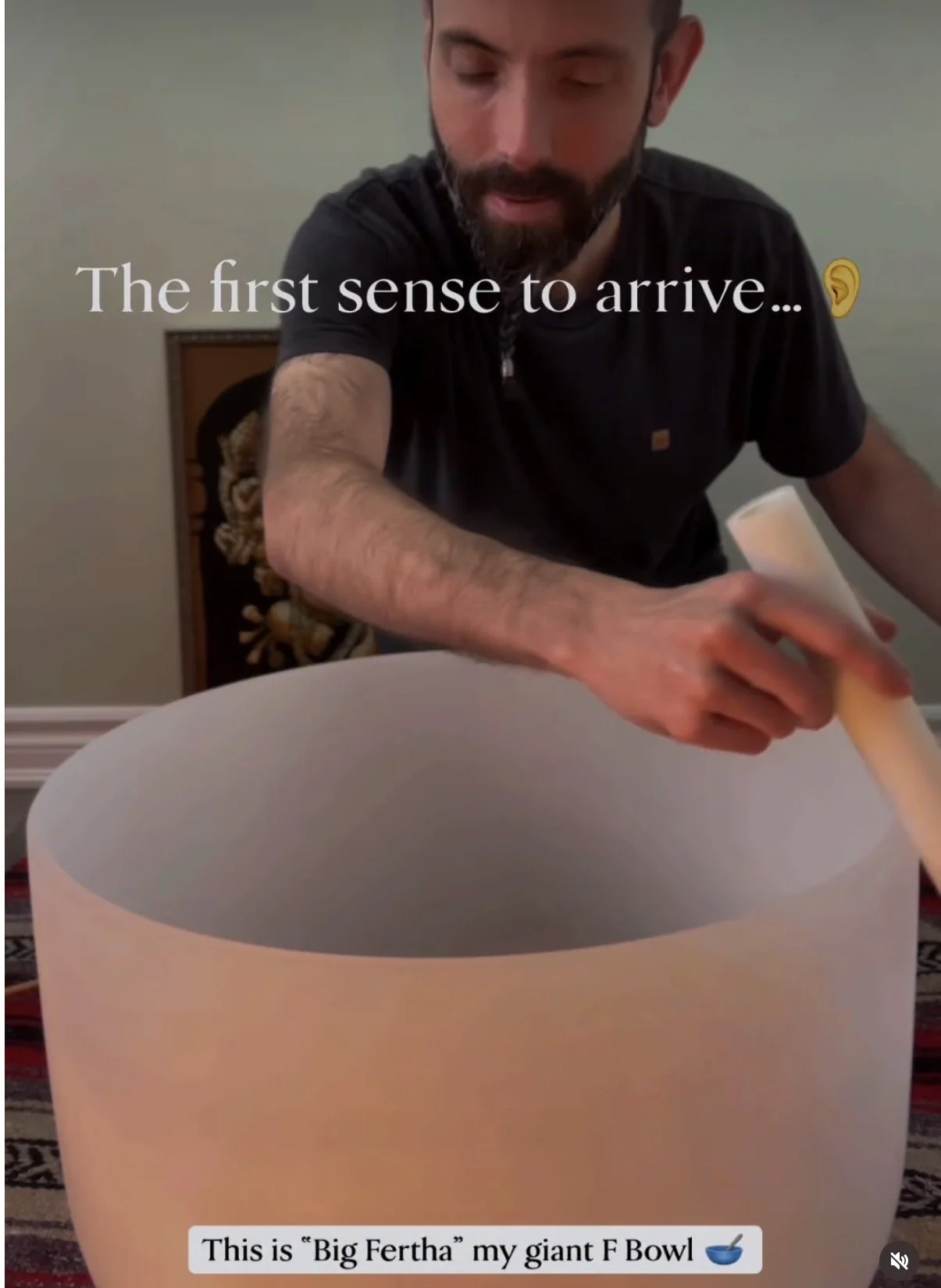 A man is playing a large crystal singing bowl, called "Big Fertha" with a wooden mallet. The text over the image says "The first sense to arrive..."