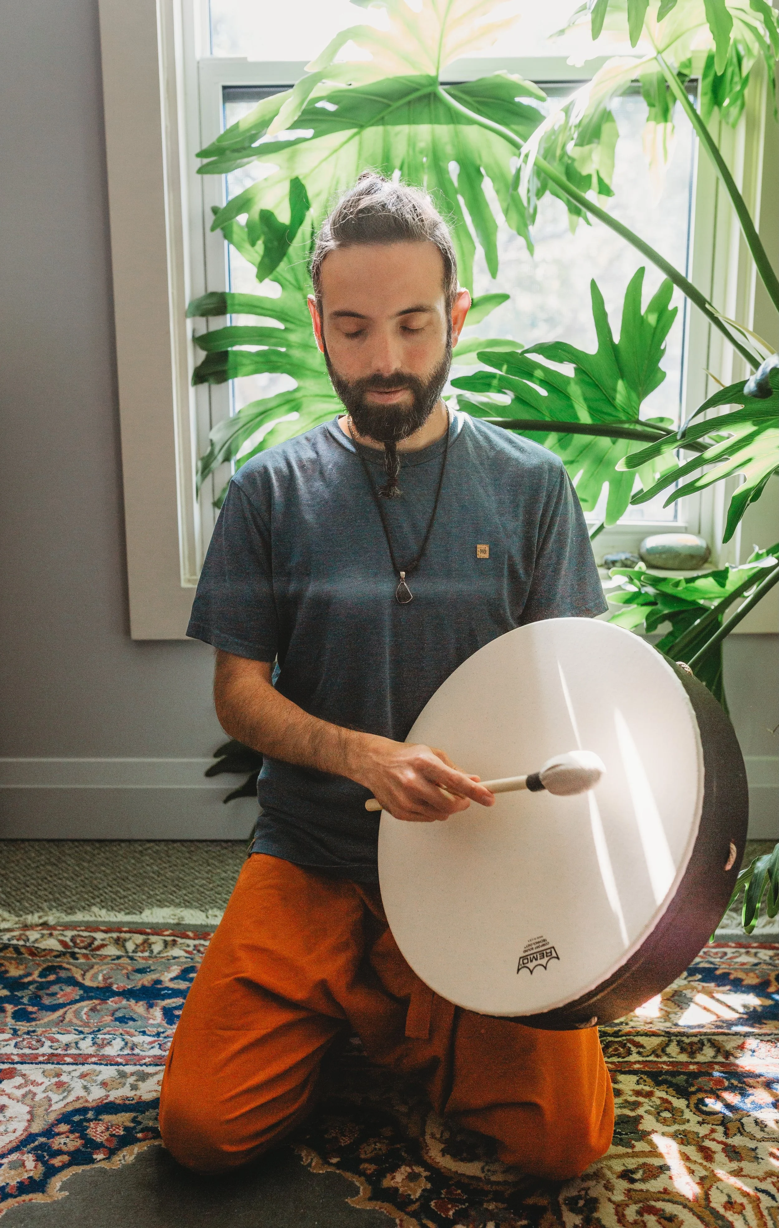 A man kneeling on a patterned rug, holding a drumstick and playing a large hand drum in a room with large green plants and a window in the background.