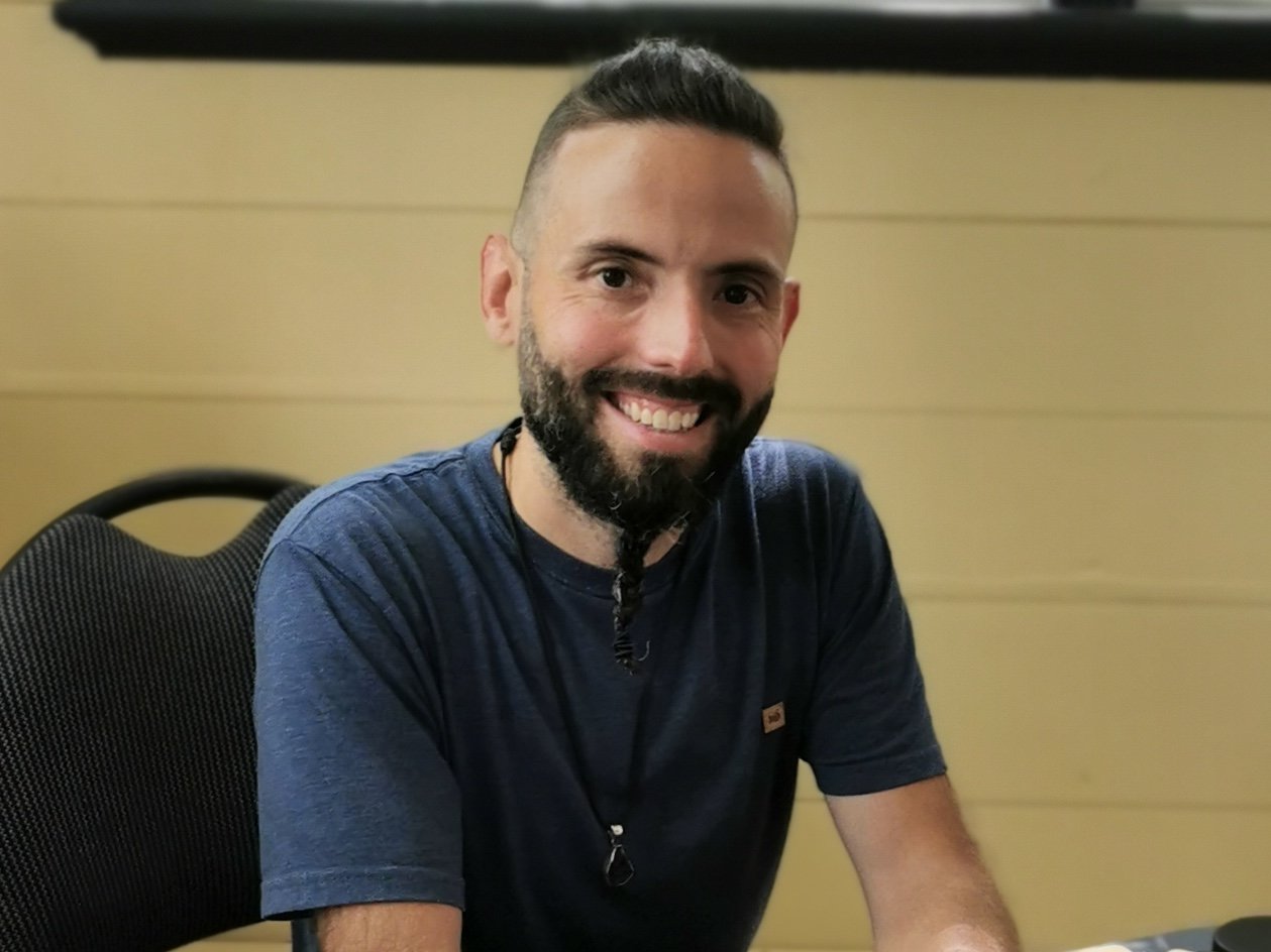 A smiling man with a beard and mustache, short dark hair, wearing a navy blue t-shirt, sitting at a table in a room with yellow wooden panel wall.