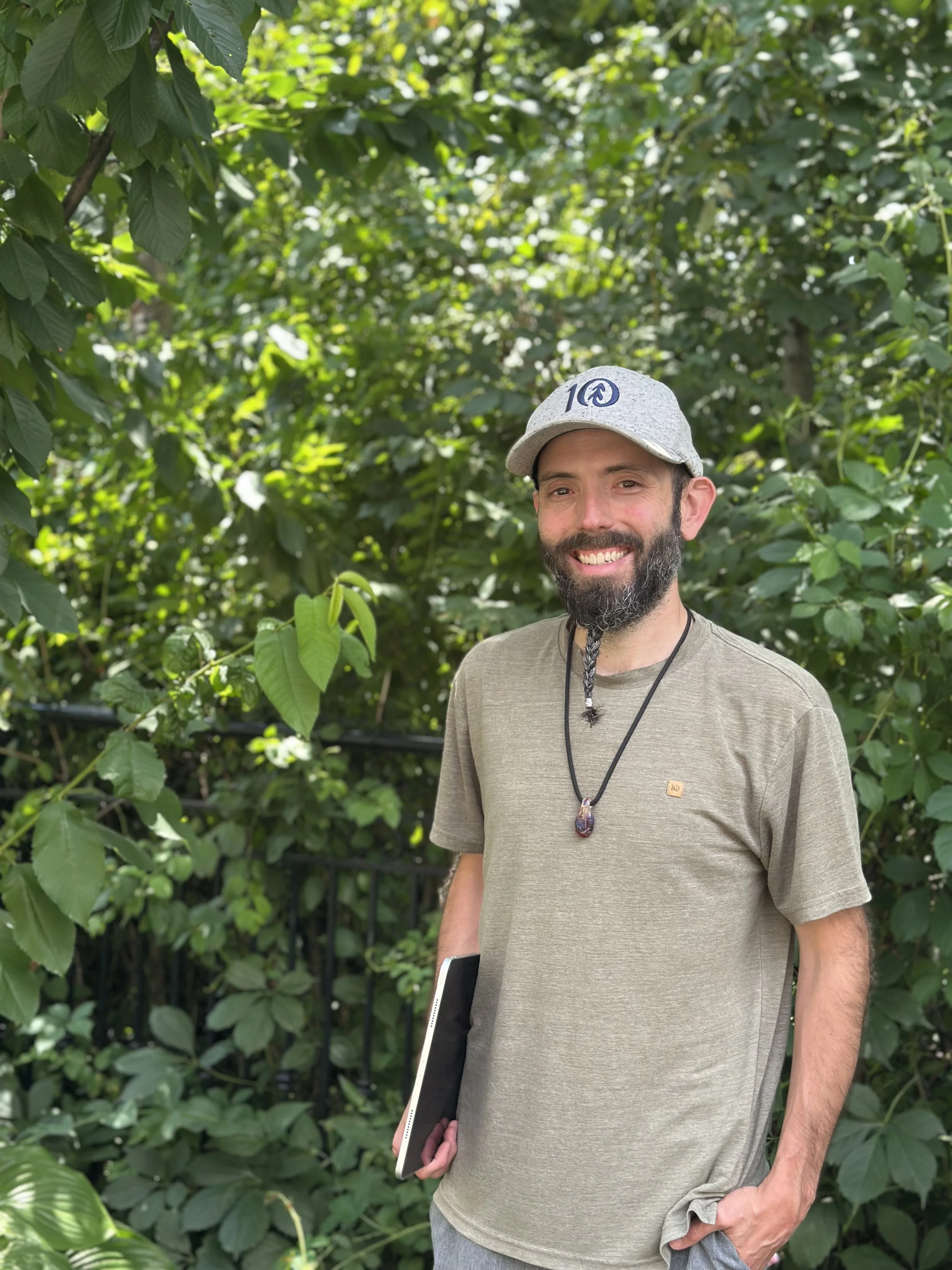 A smiling man with a beard standing outdoors in a green, leafy environment, holding a tablet or laptop, wearing a gray cap, a beige t-shirt, and a necklace.