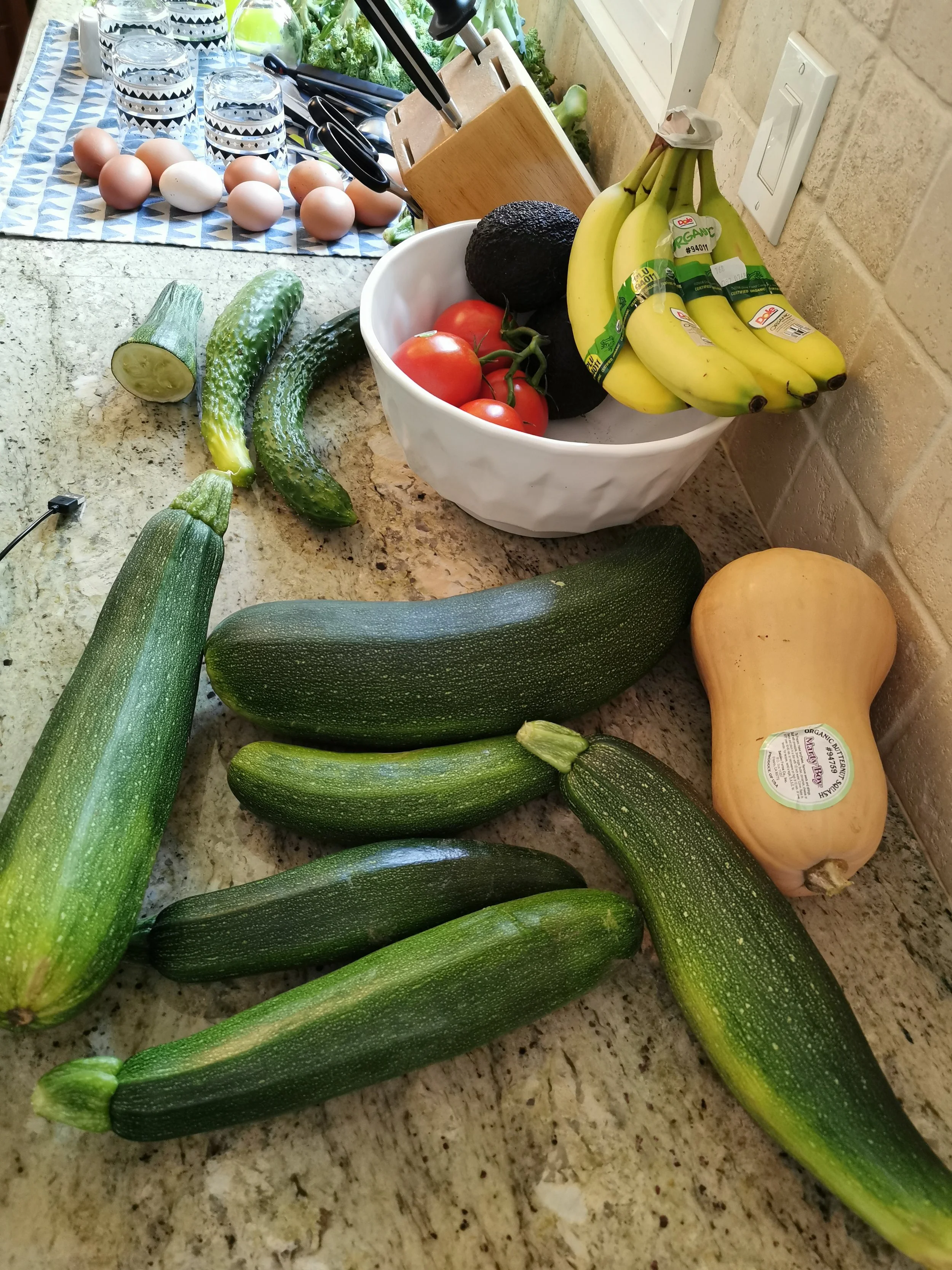 A variety of fresh vegetables including zucchini, tomatoes, avocados, and butternut squash on a kitchen countertop, with eggs and glasses of water in the background.