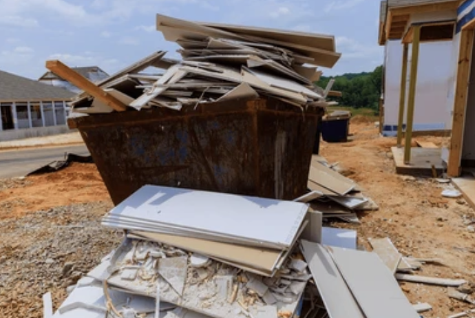 Construction debris including drywall and wood scraps in a large rusted dumpster at a building site with under-construction buildings nearby.