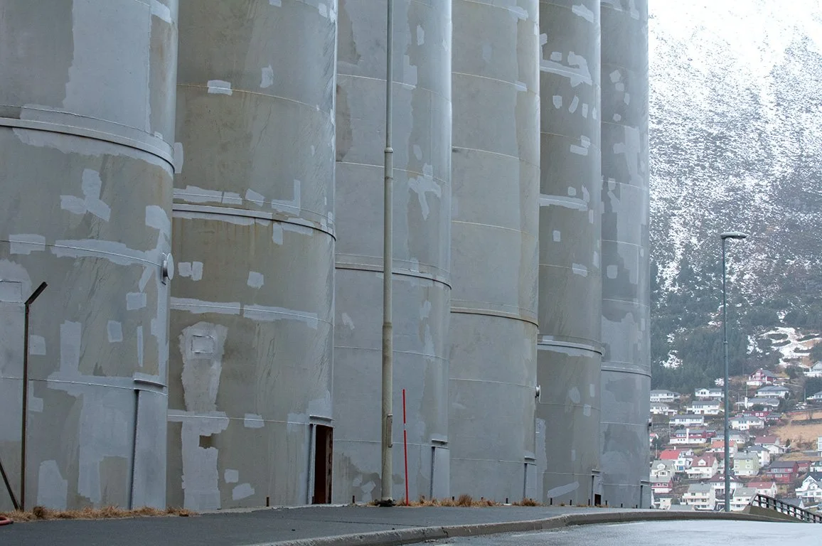 Large concrete silos with patches of repaired paint, situated on a sloped road in a hilly residential area with houses and trees in the background.