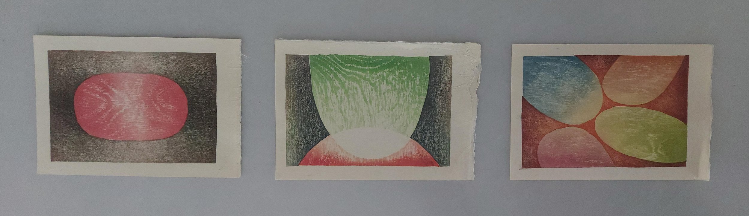 Three abstract colorful prints on paper, arranged horizontally on a gray wall. The first features a pink oval with textured details. The second has a circular shape with green, red, and black sections and textured patterns. The third displays multiple overlapping oval shapes in shades of blue, yellow, red, and pink with textured details.