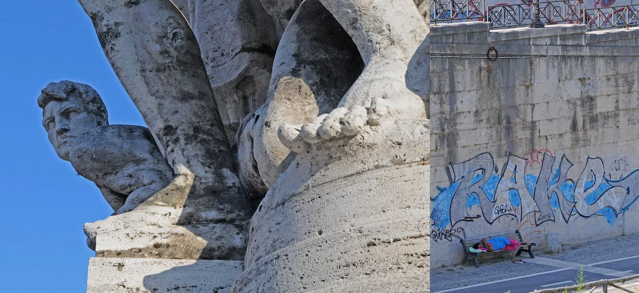 Close-up of the ancient marble sculpture part of the Winged Victory of Samothrace, showing a person's face and wing, against a clear blue sky; nearby, a person sleeps on a bench at a graffitied wall.