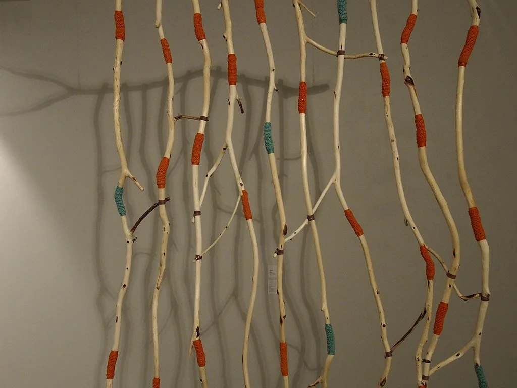 Wall hanging art piece made of woven wooden sticks with orange and green wrapped sections, casting shadows on a gray wall.