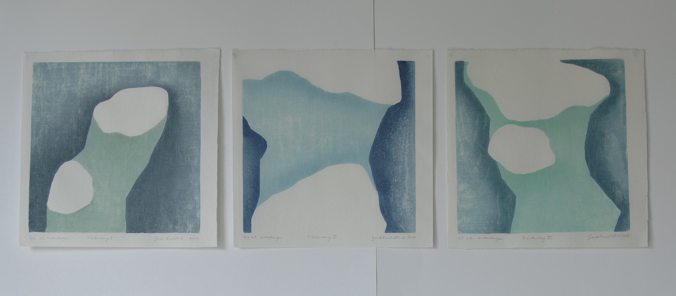 Three abstract landscape paintings featuring blue and green hues, mounted side by side on a white wall.