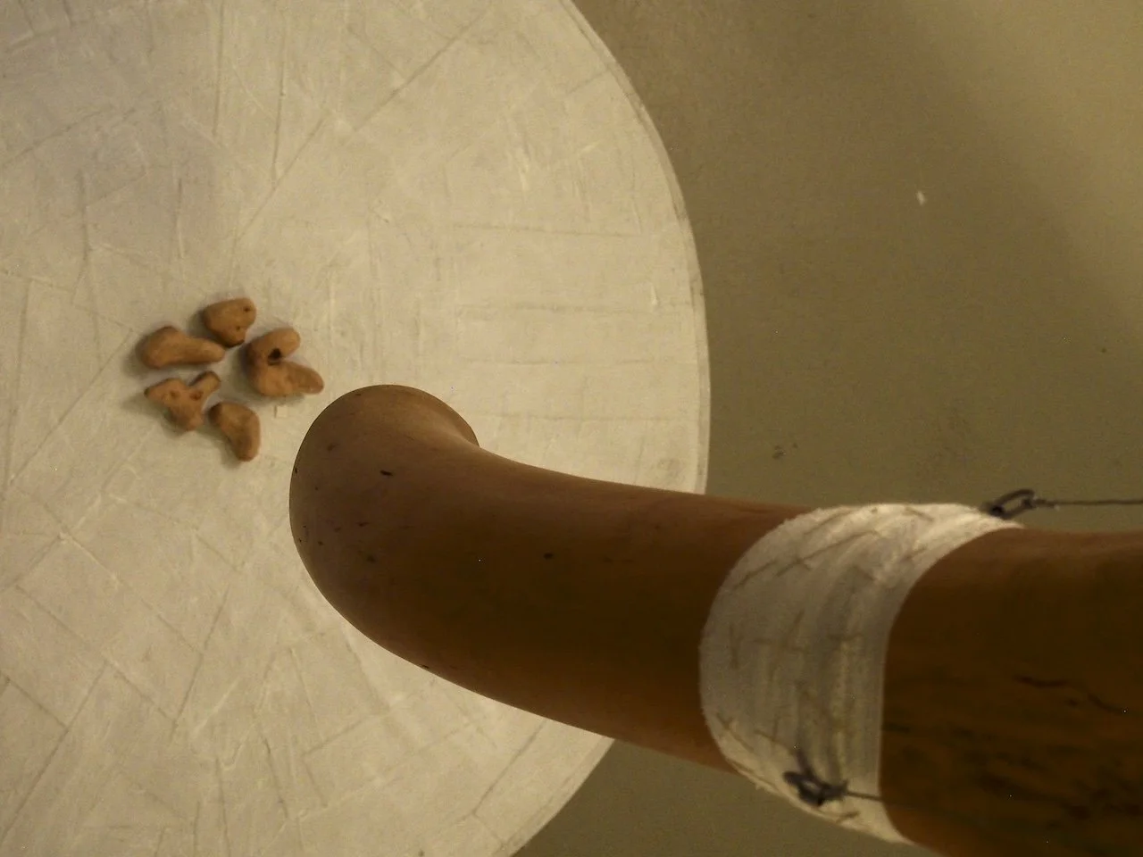 A wooden table leg with a white cloth wristband and a string, viewed from below, with scattered snack crackers on a white tiled floor.