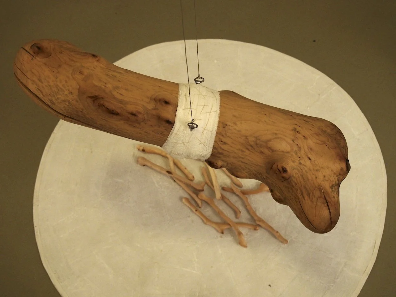 A sculpture of a dog made from a large piece of wood, mounted on a round white base with some twigs or roots at the bottom.