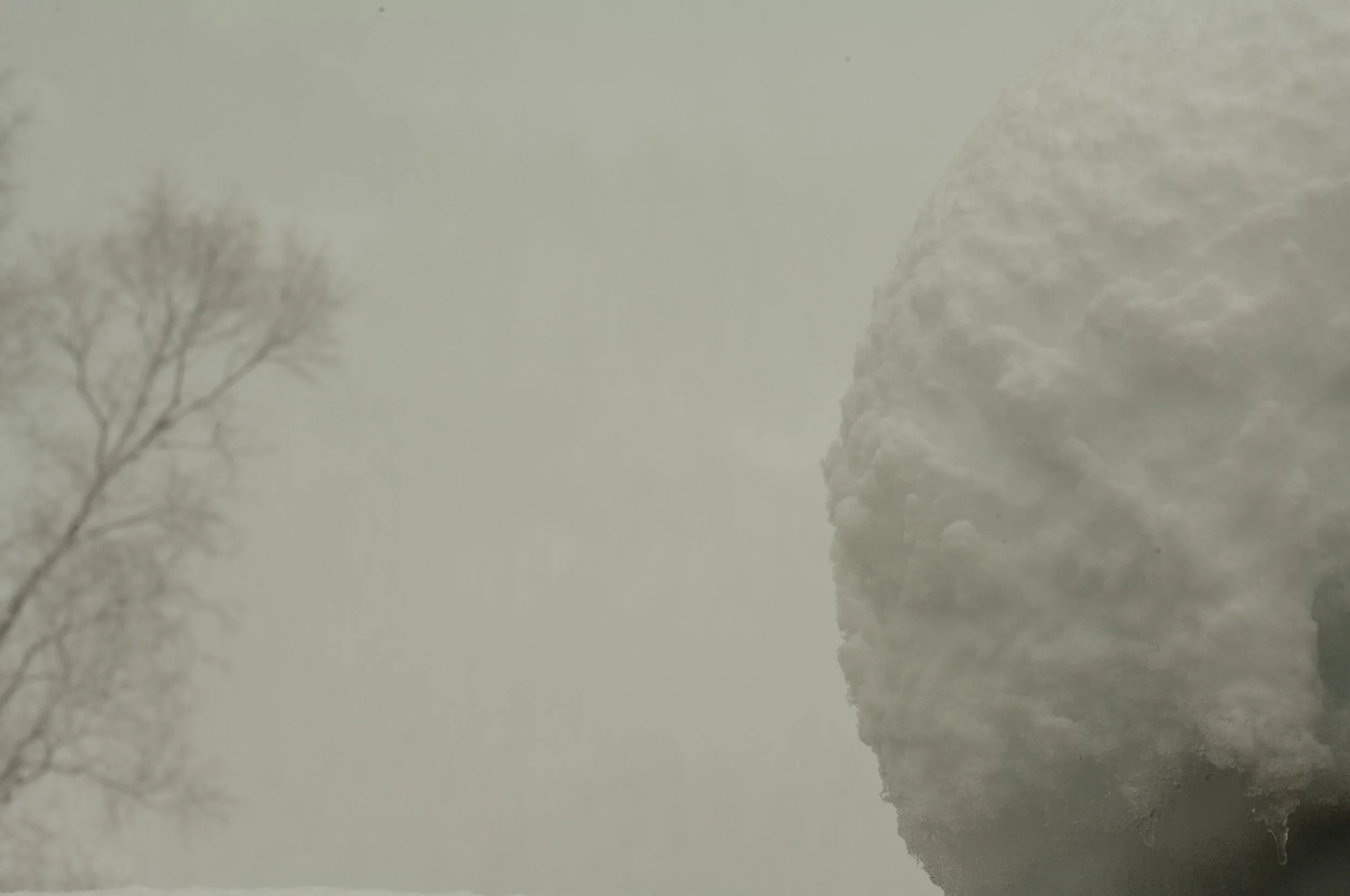 A foggy winter scene with a leafless tree on the left and a large snow-covered rock or structure on the right.