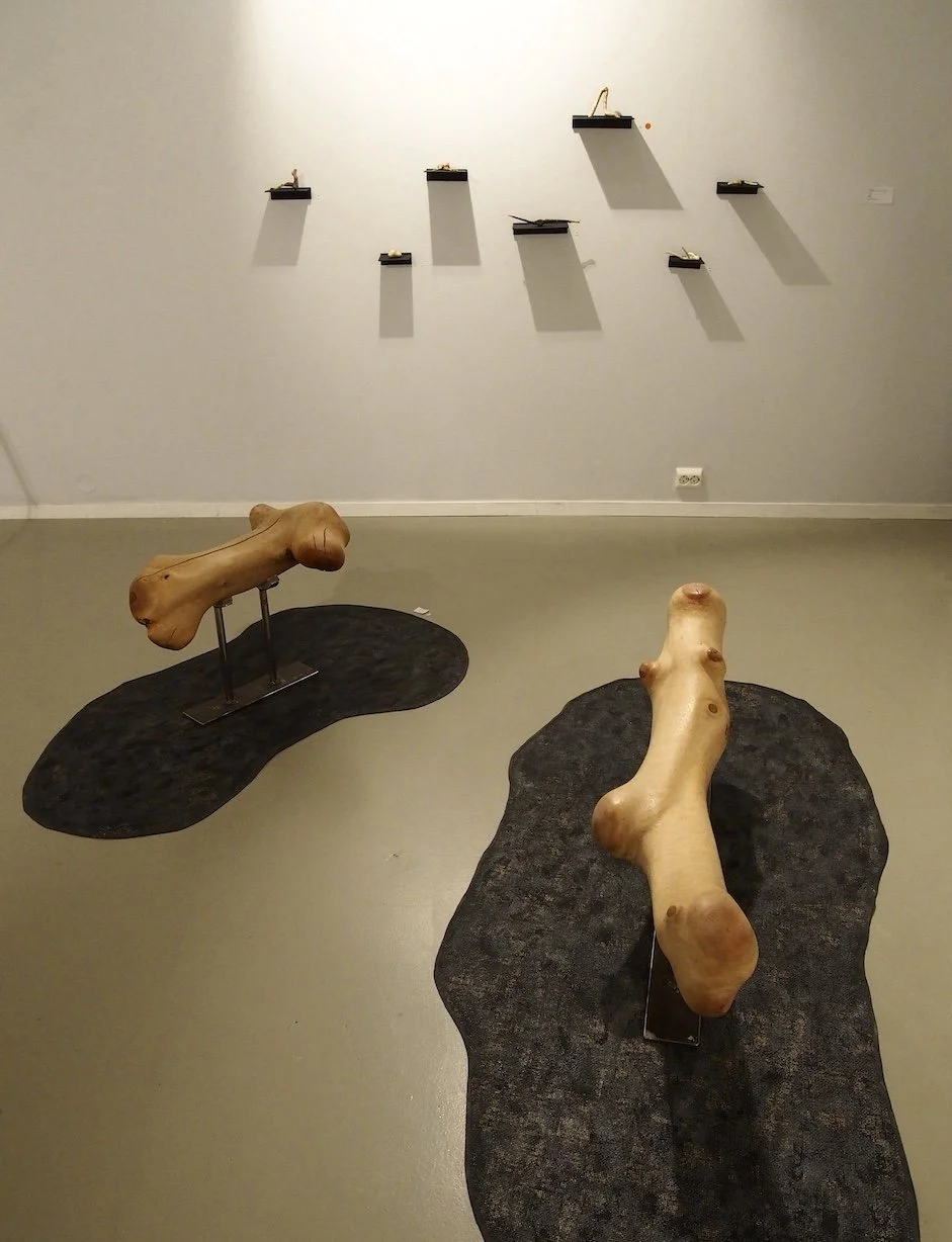 Contemporary art gallery with two wooden sculptures resembling abstracted human torsos on irregularly shaped black bases, and small wall-mounted sculptures on floating shelves along the white gallery wall.