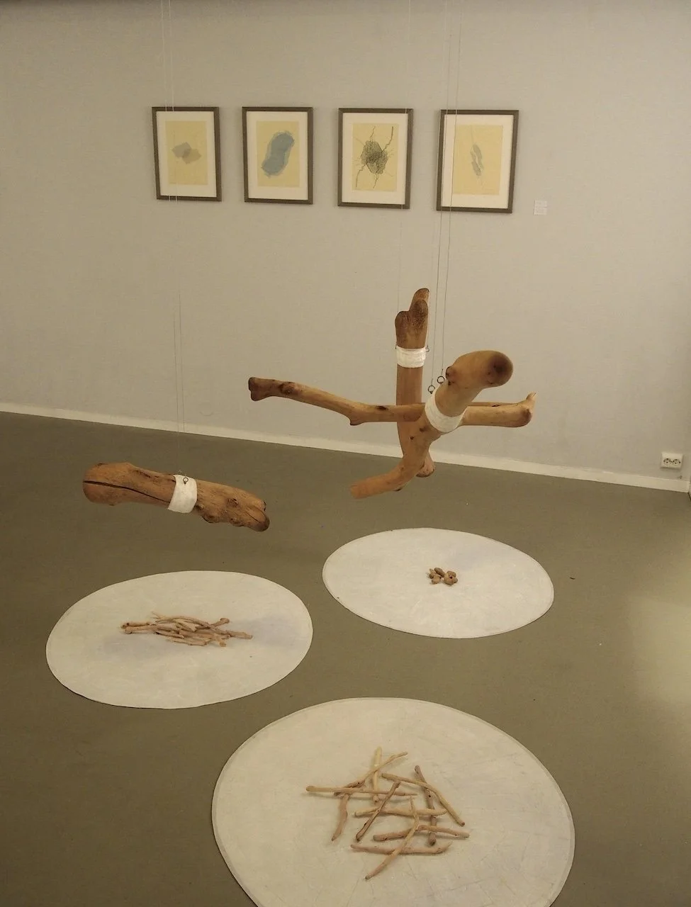 Art installation featuring a suspended wooden sculpture of a child's figure with bandages, surrounded by three circular rugs on the floor with wooden sticks and pieces arranged in different patterns, and framed abstract artwork on the wall.
