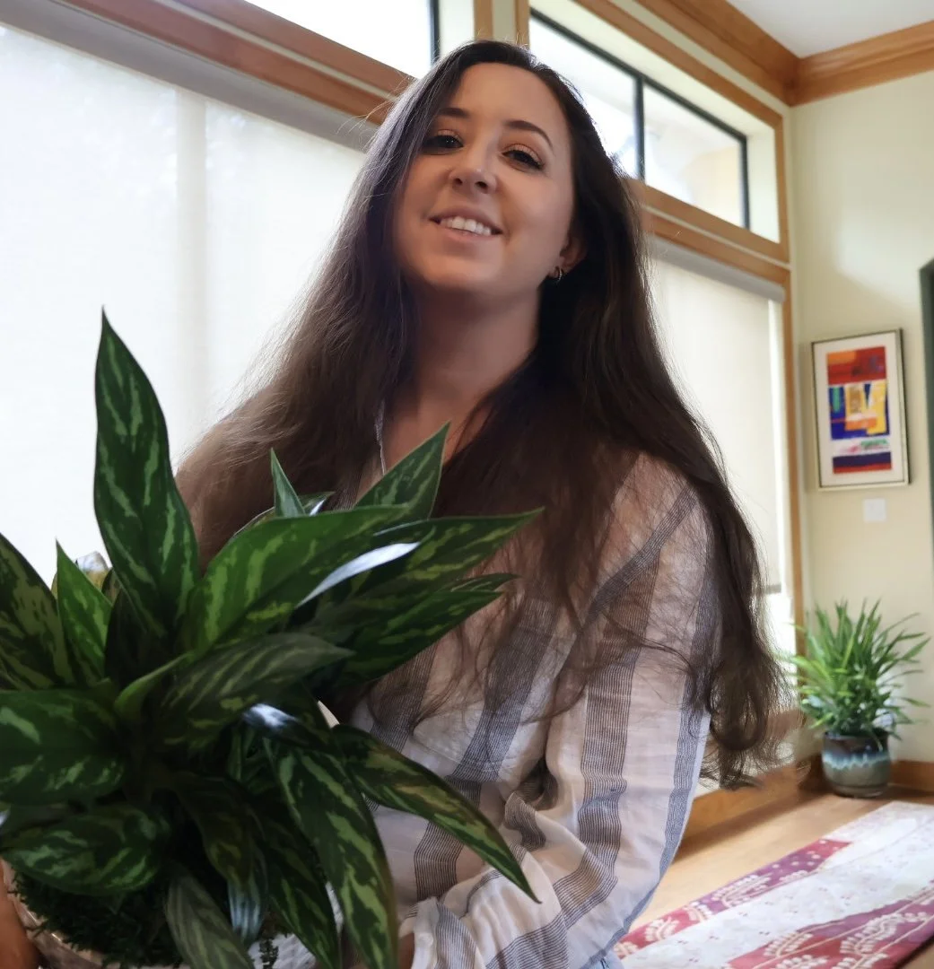 A woman with long dark hair smiling and holding a large green leafy houseplant in a well-lit room with large windows, wooden accents, and colorful artwork.