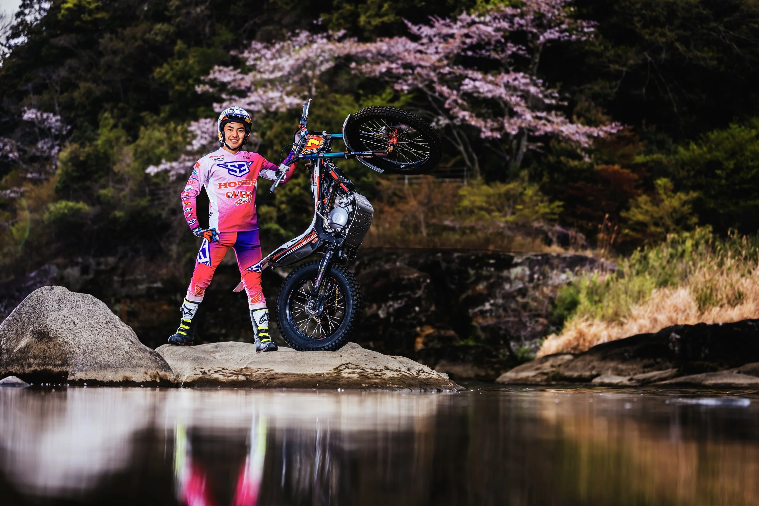 女性がオフロードバイクで逆立ちをしながら自然の中の岩の上に立っている。背景にはピンクの桜の木と緑の木々が見える。