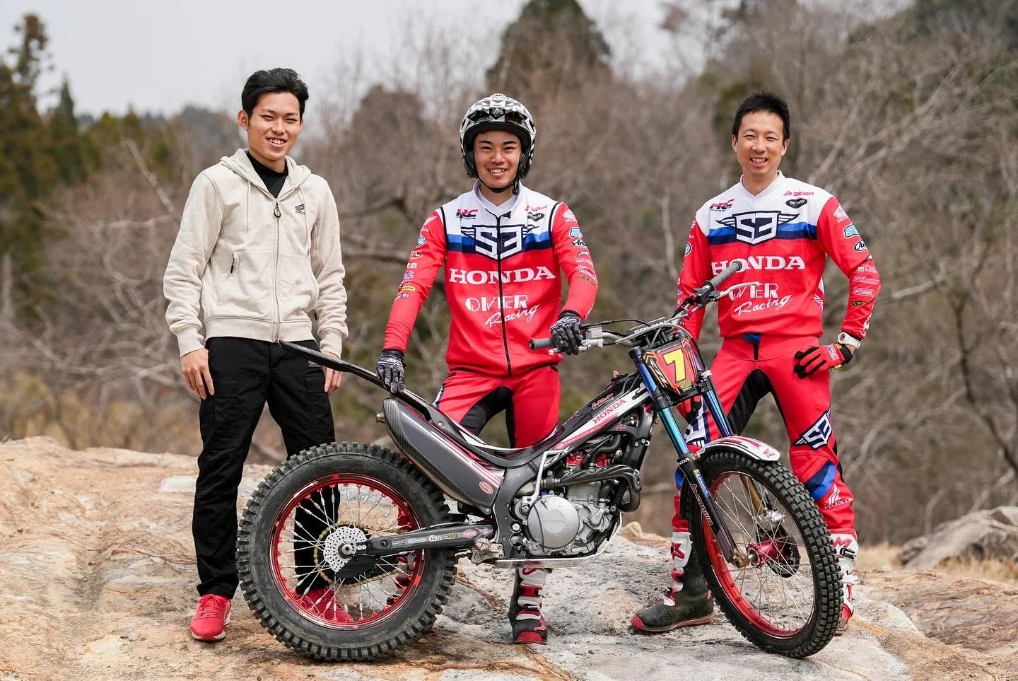 4人の若者、うち2人はマウンテンバイクのライダー、残りは観客またはサポートスタッフ、屋外の岩場で、背景は木々や山。