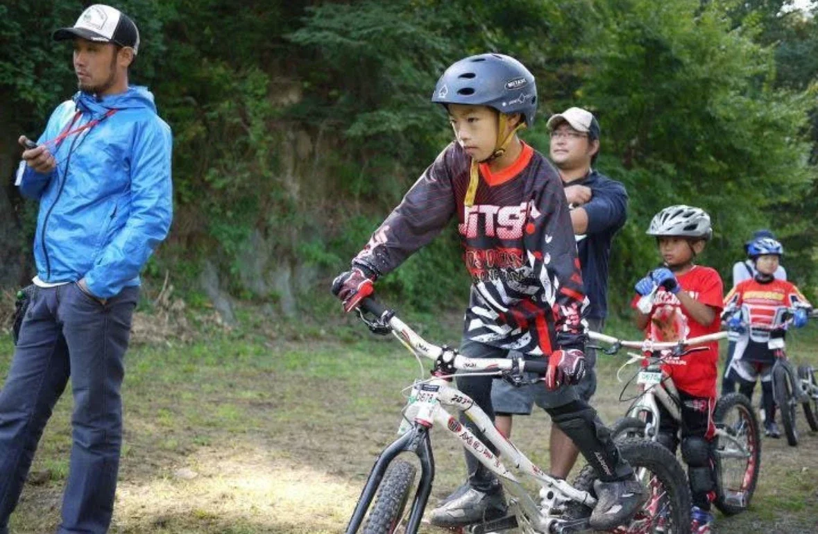 子供たちがサイクリングを楽しんでいる風景。少年はヘルメットをかぶり、自転車に乗っている。大人が見守っている。周囲は自然豊かな公園のような場所。