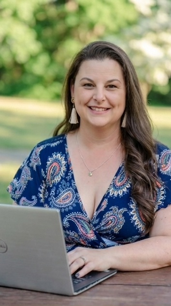 Daria Beddard with long brown hair, wearing a blue patterned blouse and earrings, sitting outdoors at a table with a laptop, smiling at the camera with a background of trees and greenery.