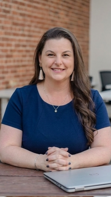 Daria Beddard with long brown hair, wearing a blue top, sitting at a table with a closed laptop in front of her, outdoors in front of a brick wall.