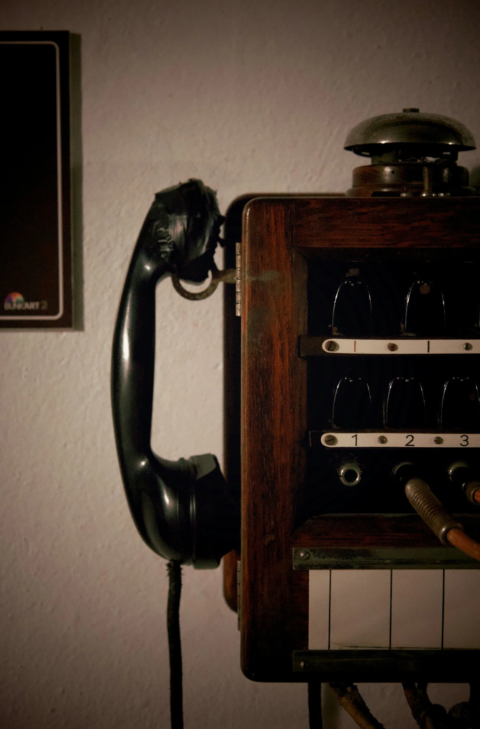 An old-fashioned black rotary wall phone mounted on a wall next to a wooden radio or electronic device with tubes and control dials.