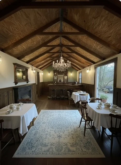 Dining room with wooden vaulted ceiling, chandelier, fireplace, and large windows, set up for a meal with round tables and chairs.