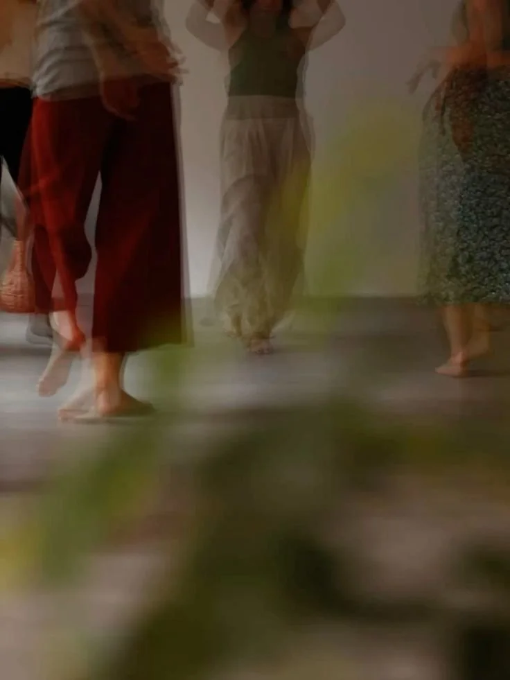 Multiple people barefoot, wearing skirts and pants, standing indoors on a wooden floor.
