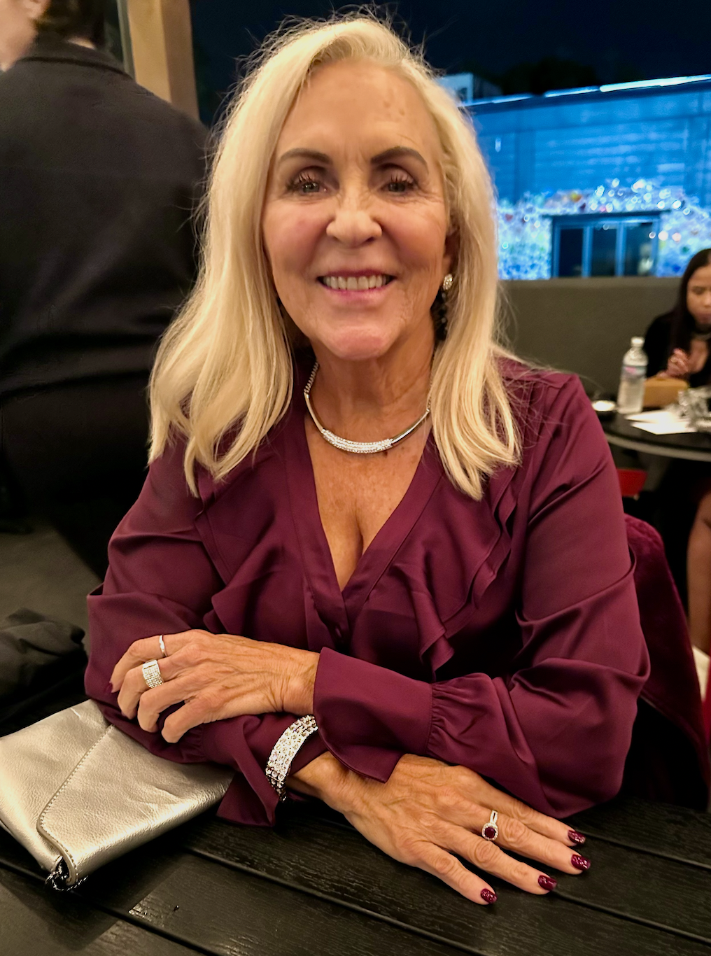 A smiling woman with long blond hair sitting at a table in a restaurant, wearing a burgundy blouse, jewelry, and a ring, with a silver purse on the table.