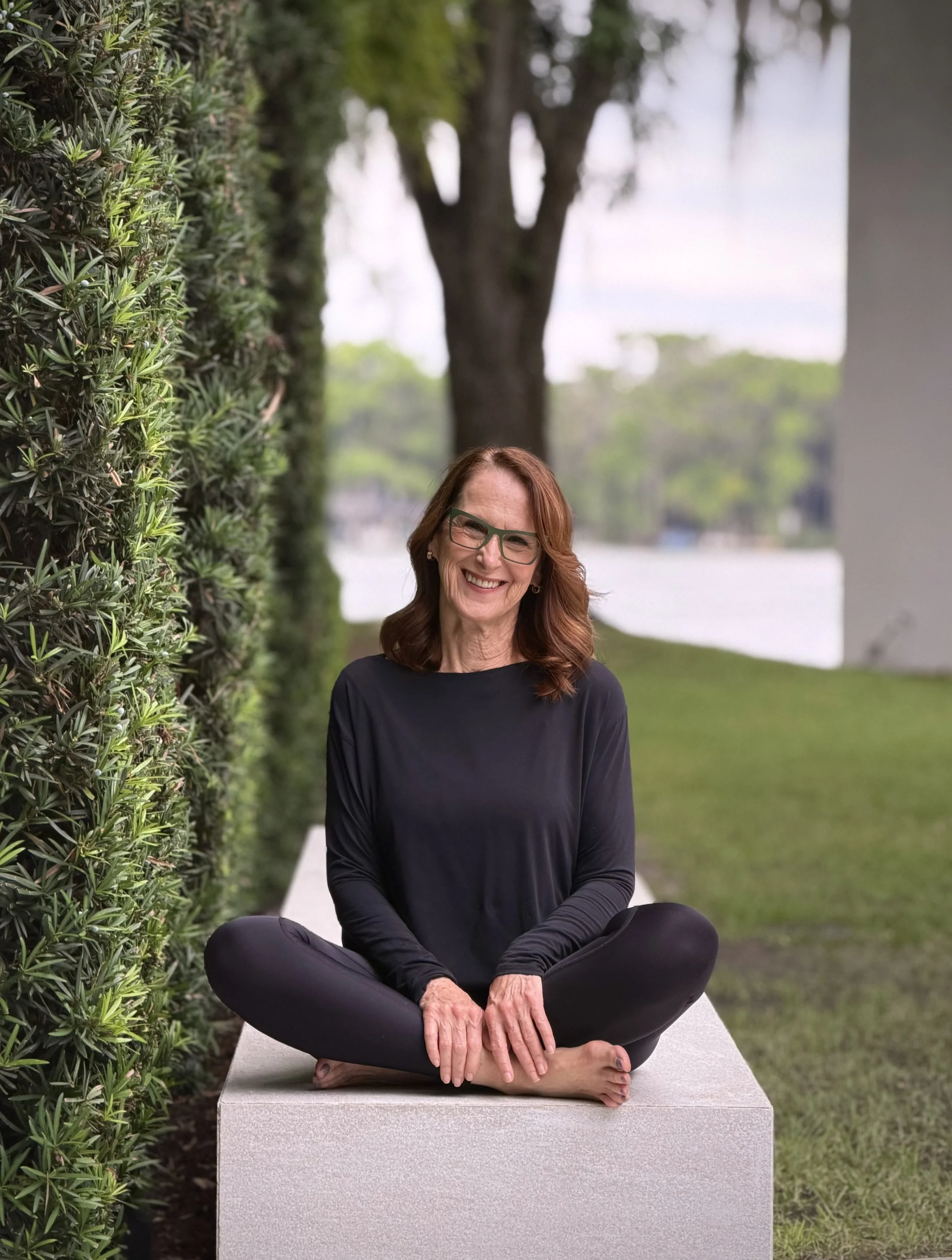 A smiling woman with red hair, glasses, and a black long sleeve shirt sitting cross-legged on a bench outdoors, practicing yoga or meditation, with a hedge and tree in the background.