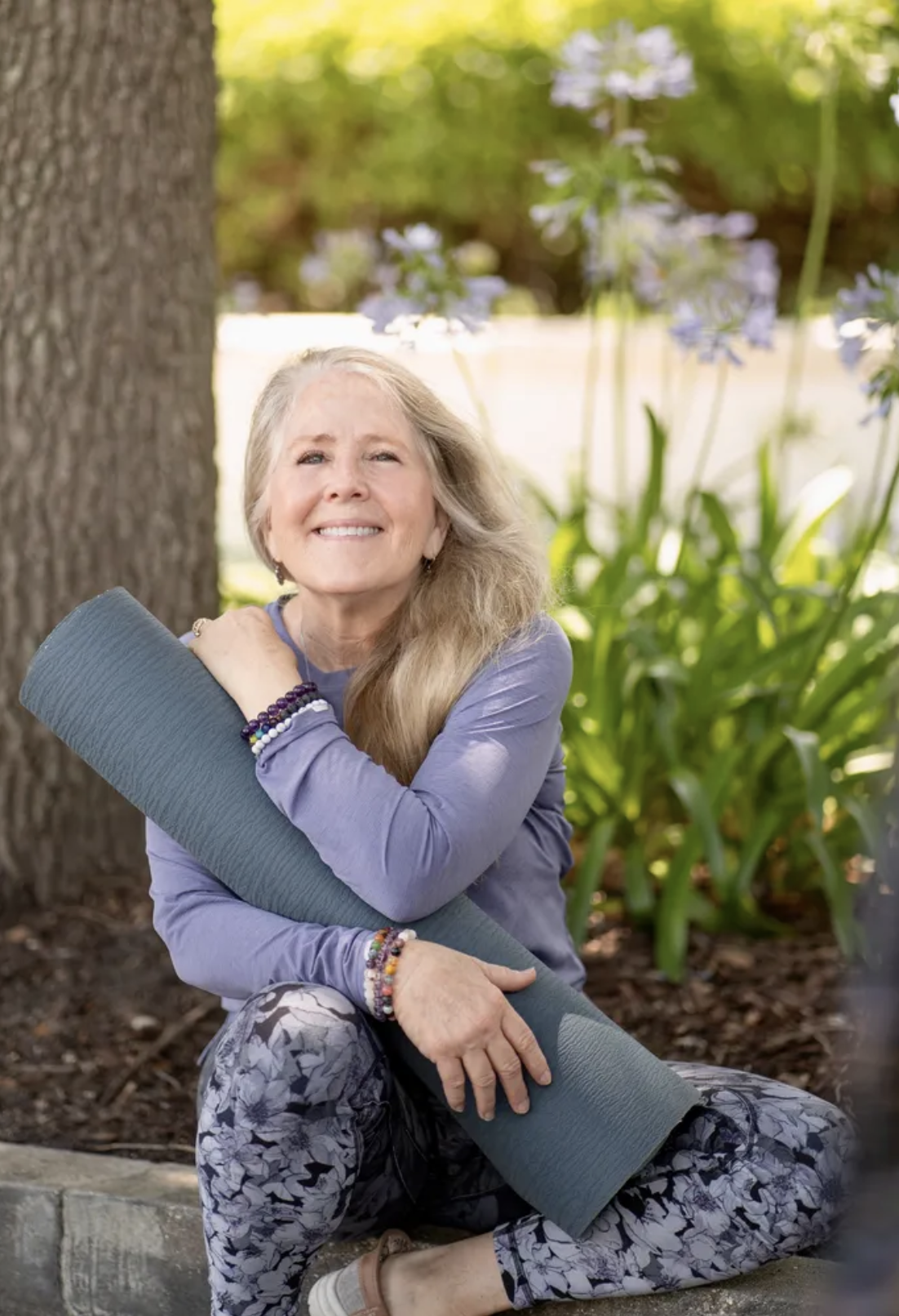 Woman with long blonde hair, smiling, comforted by a yoga mat, sitting outdoors near a tree and green foliage, dressed in purple long-sleeve shirt and patterned leggings.